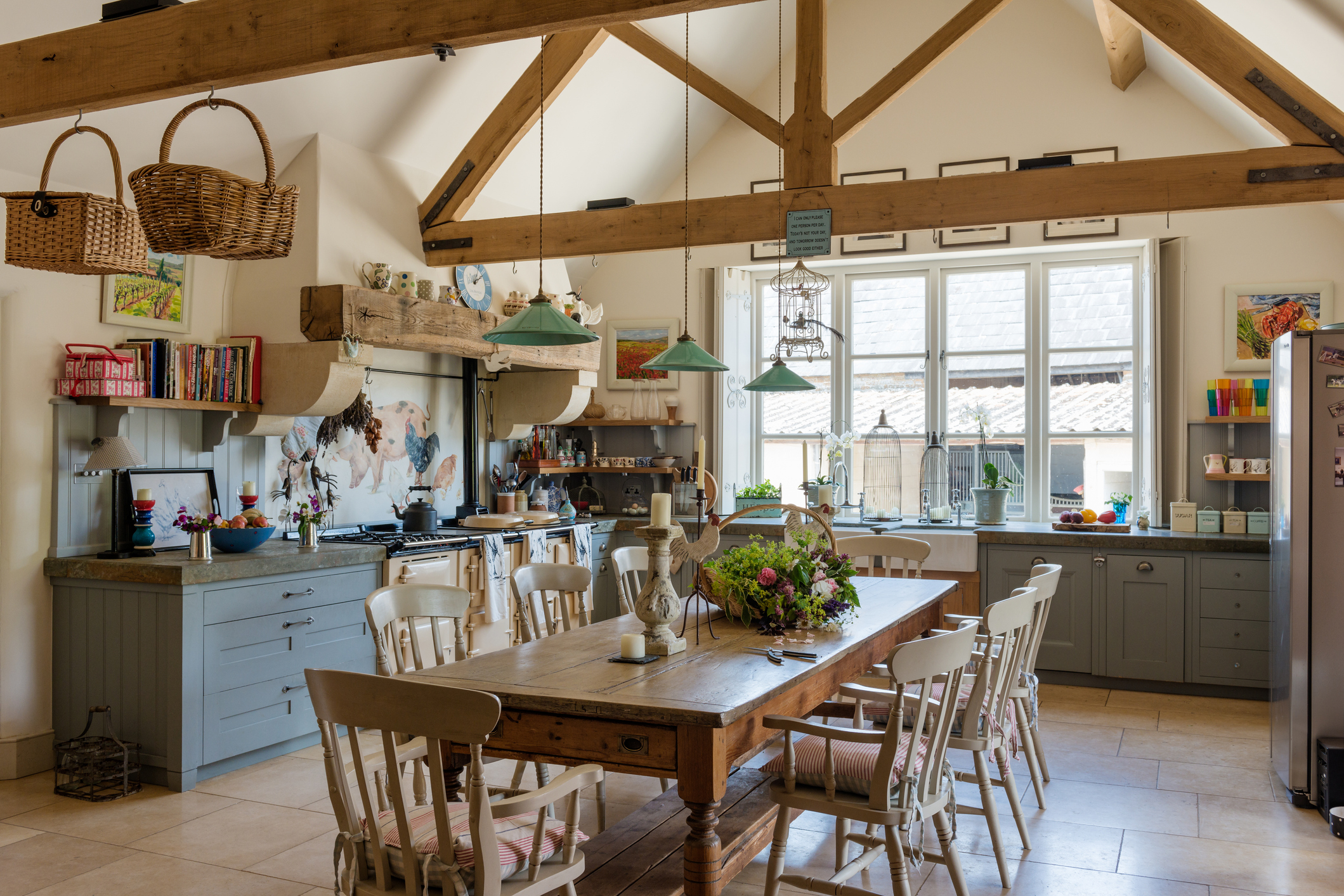 Spacious rustic kitchen with wooden beams, large dining table, chairs, hanging light fixtures, and an assortment of shelves, cabinets, and decor