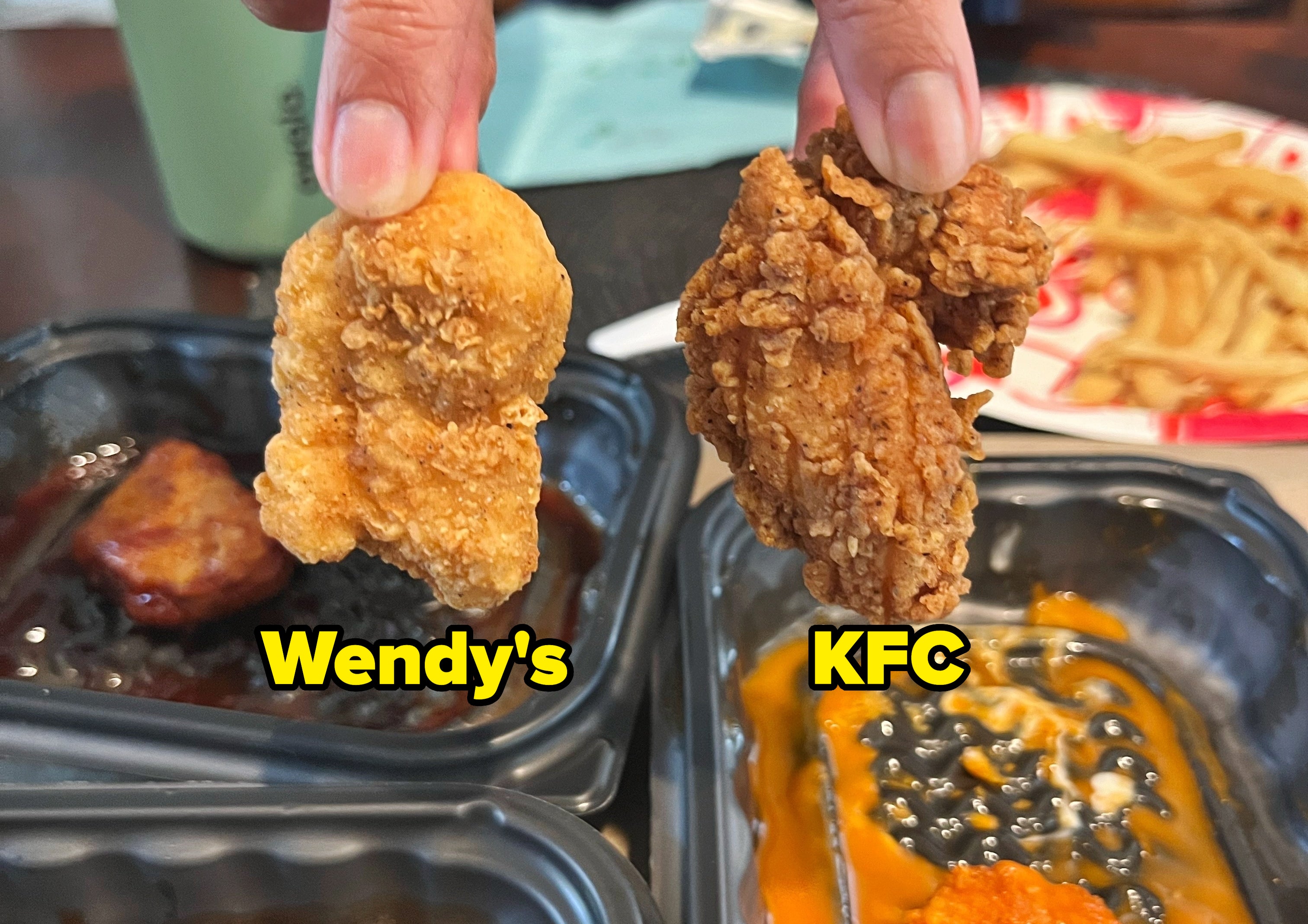 Person holding a chicken nugget and a fried chicken piece above dipping sauces, served with fries in a food container