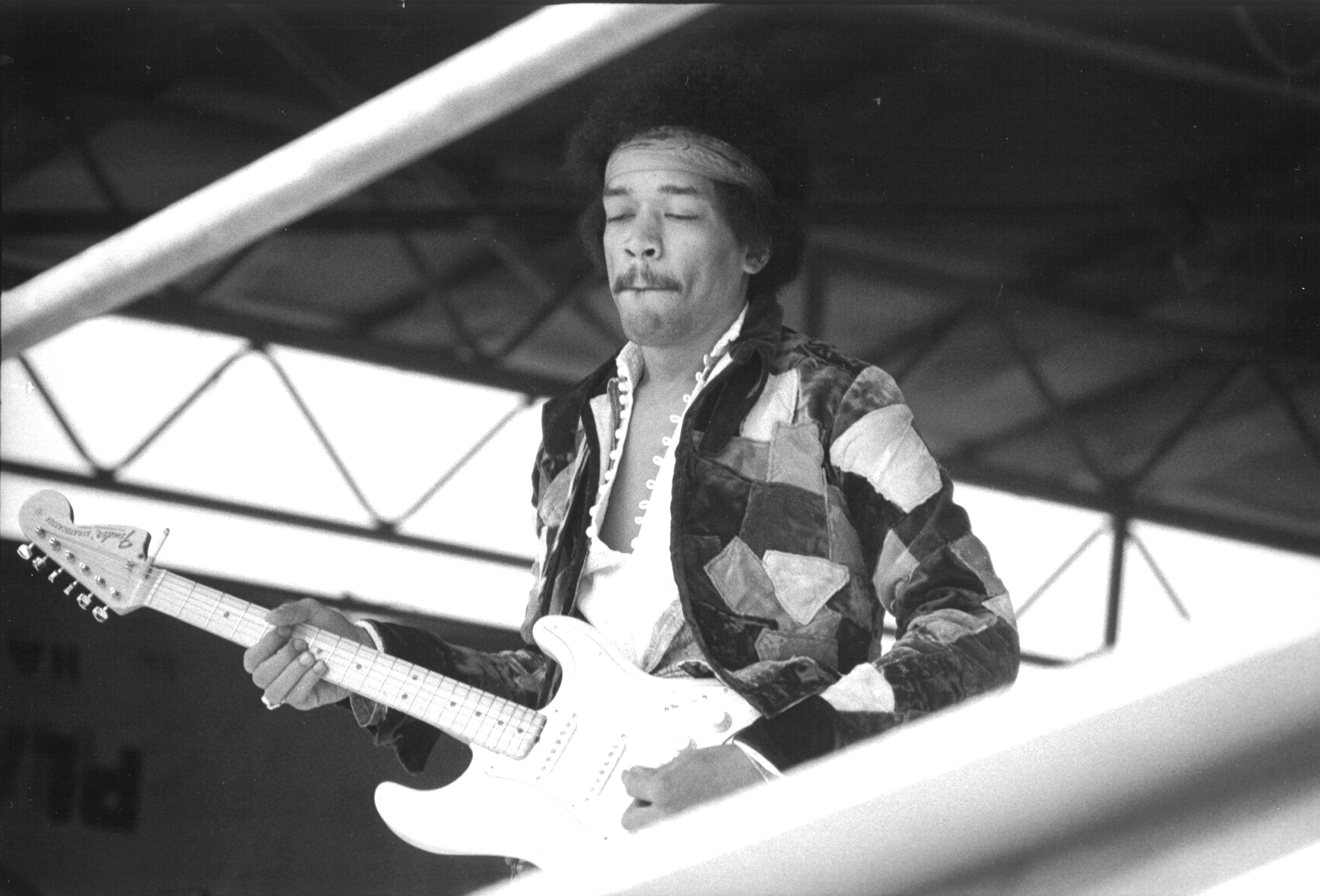 Jimi Hendrix performing on stage playing an electric guitar, wearing a patterned jacket and headband