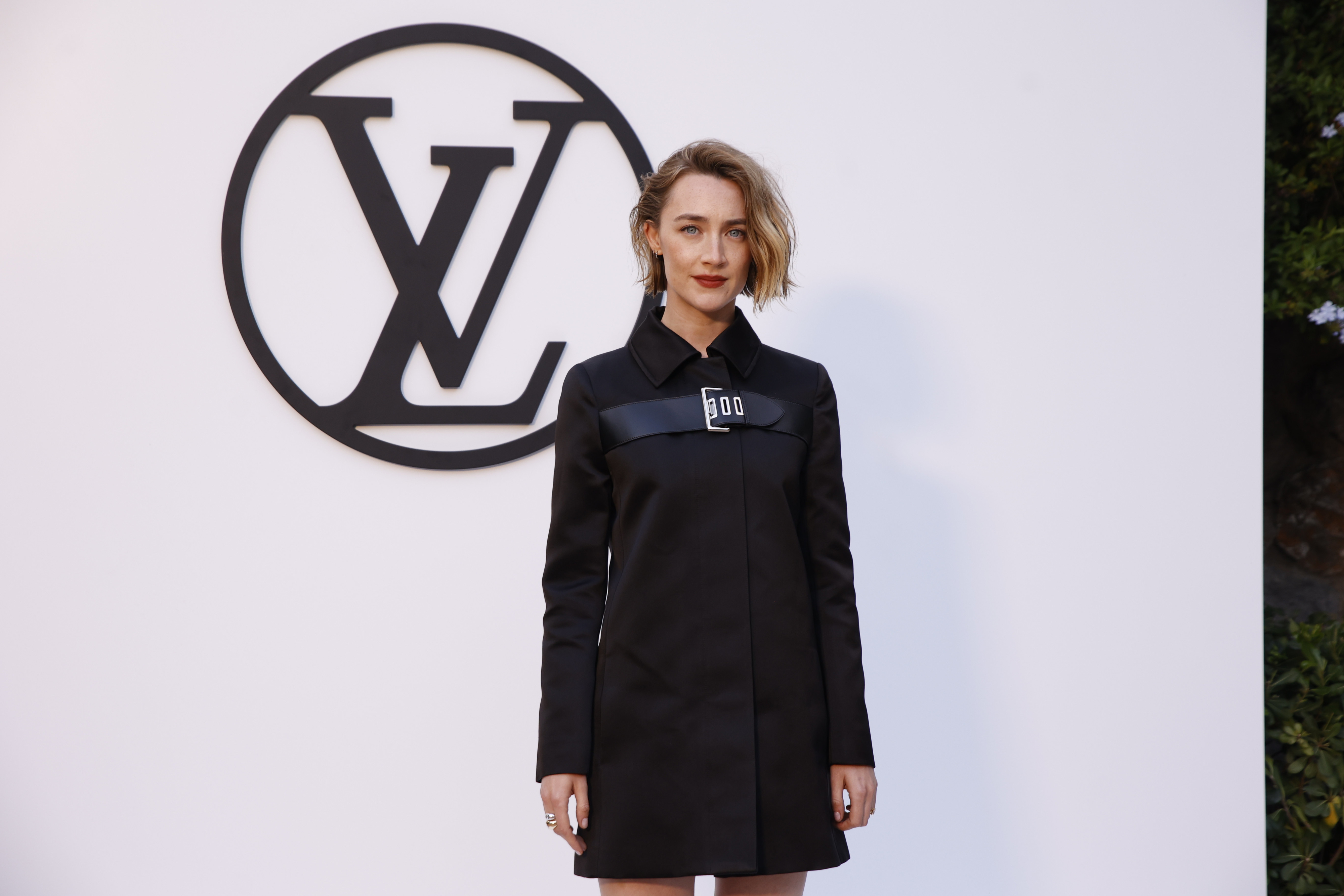 Saoirse Ronan poses on the red carpet in a stylish black dress coat in front of a Louis Vuitton logo backdrop