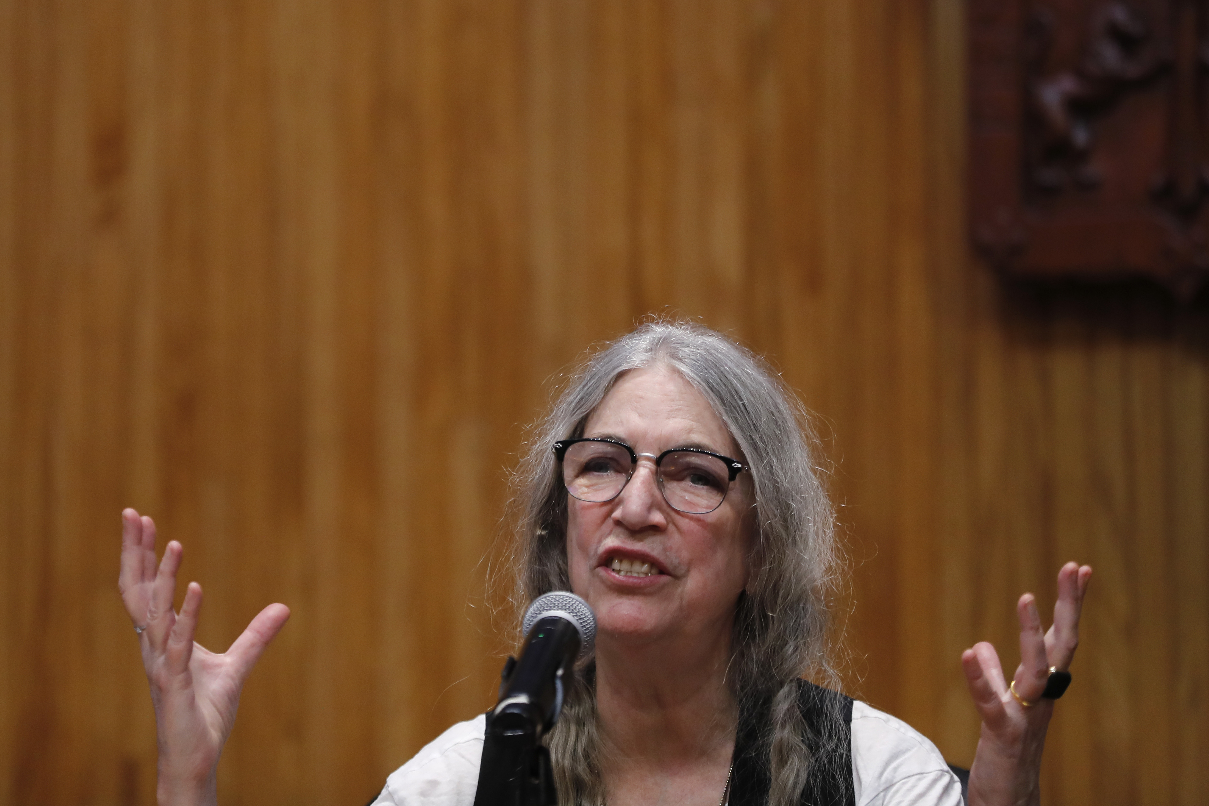 Patti Smith, wearing glasses and a casual outfit, speaks passionately at a microphone with hands raised against a wooden background