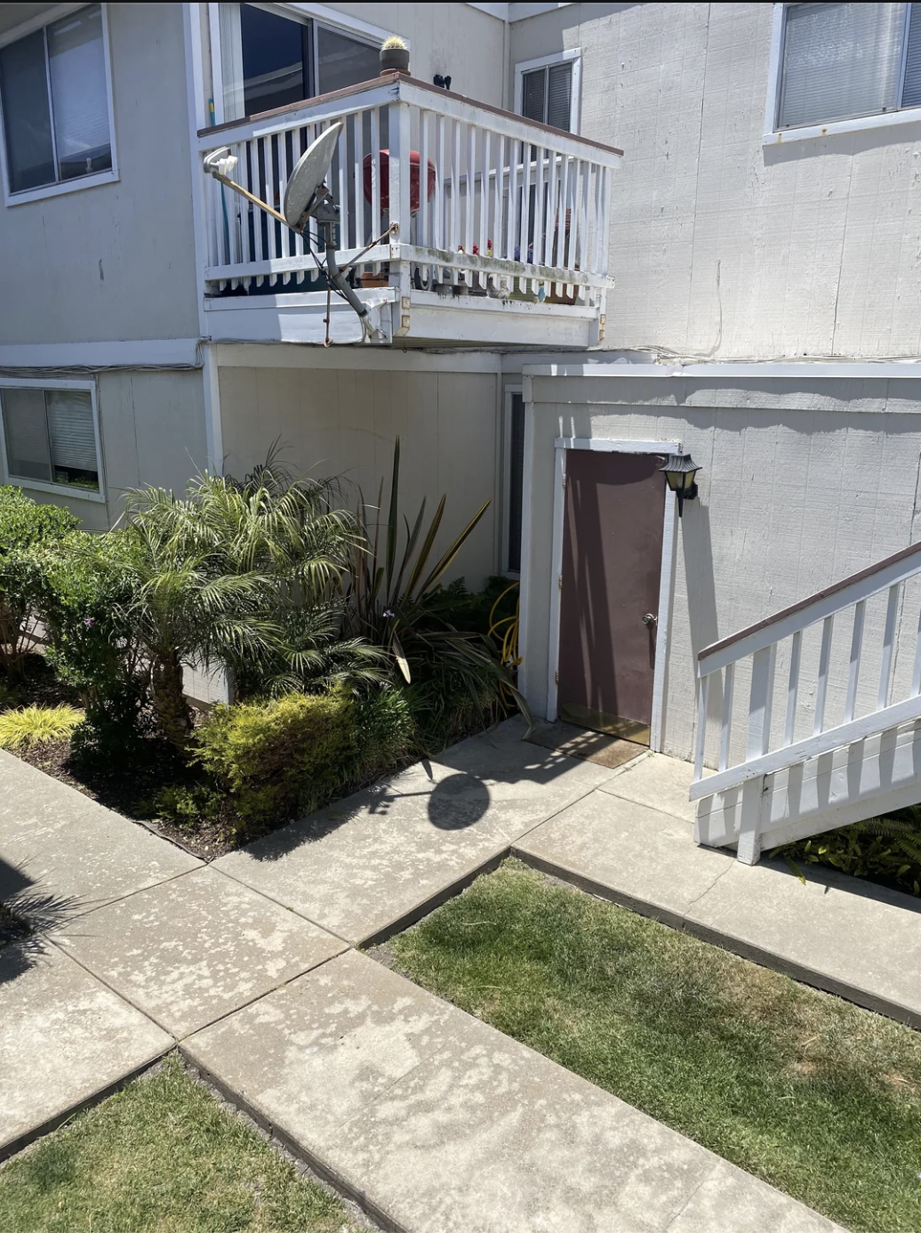 A person is standing on the second-floor balcony of an apartment building, looking over the railing. The ground level features concrete pathways and greenery