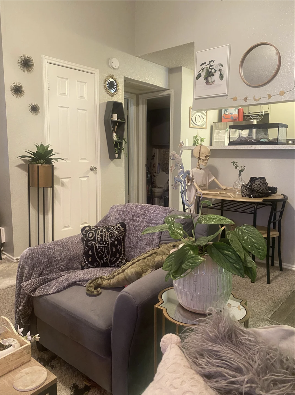 Cozy living room with potted plants, a gray armchair with a blanket, decorative wall art, and a crocodile plush toy