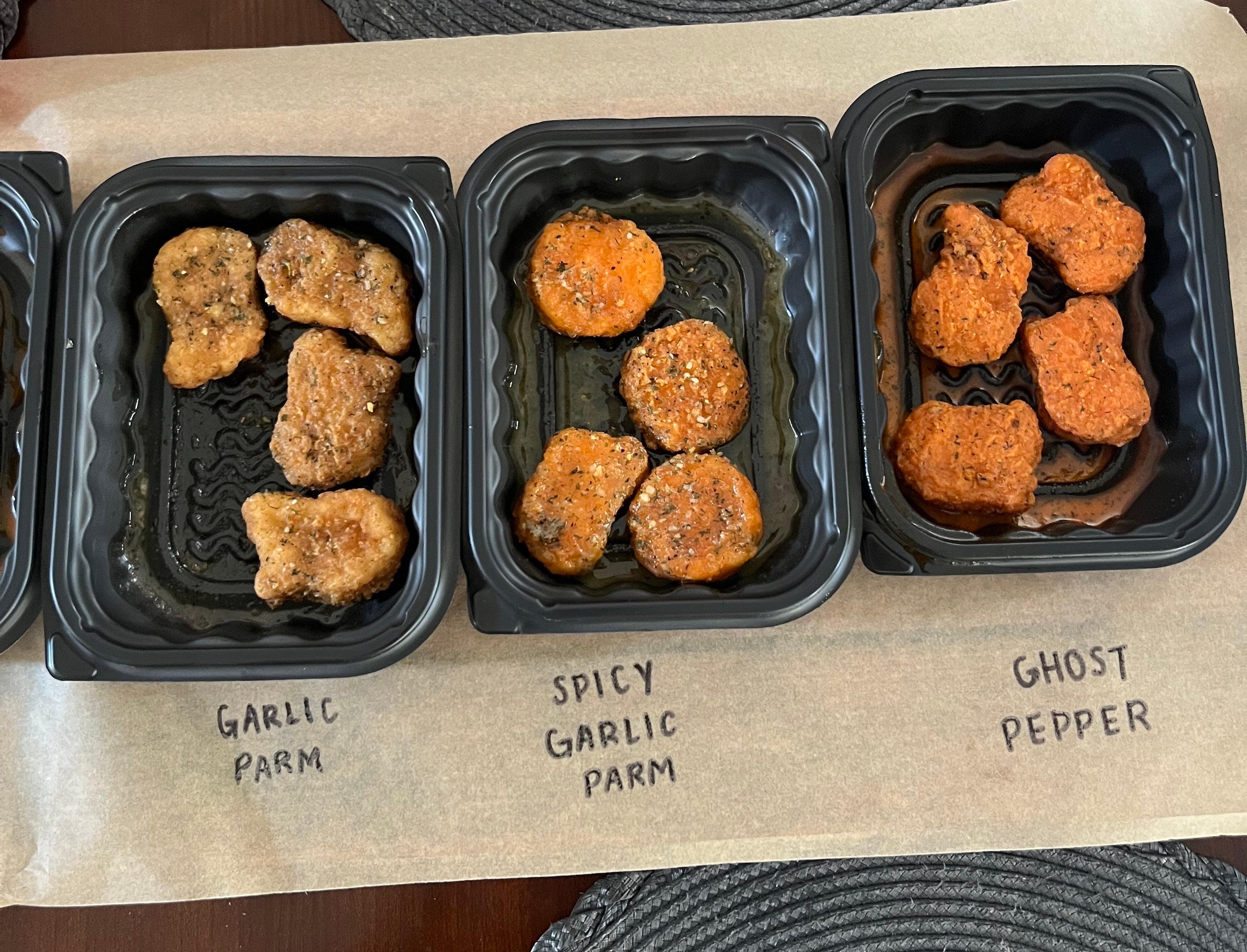 Three trays of chicken wings labeled &#x27;Garlic Parm,&#x27; &#x27;Spicy Garlic Parm,&#x27; and &#x27;Ghost Pepper&#x27; are displayed on a table