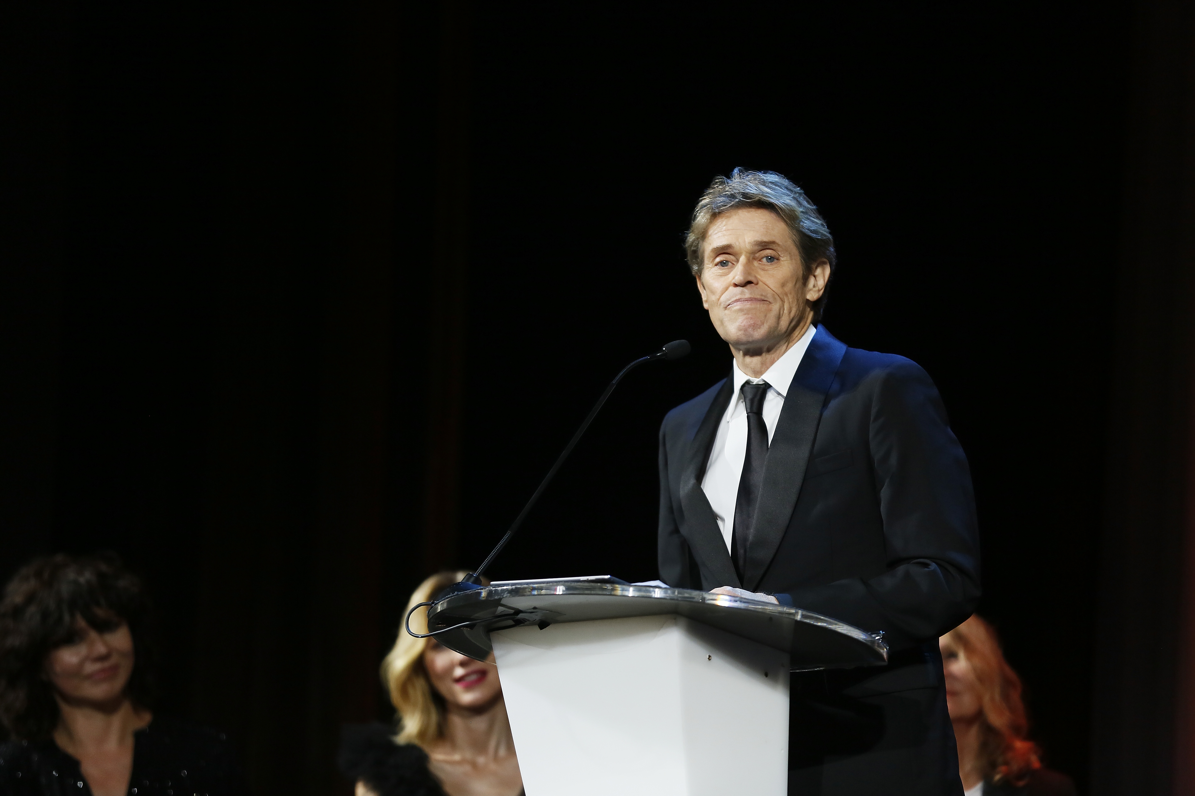 Willem Dafoe speaks at a podium, wearing a black suit with a white shirt and black tie. People listen in the background