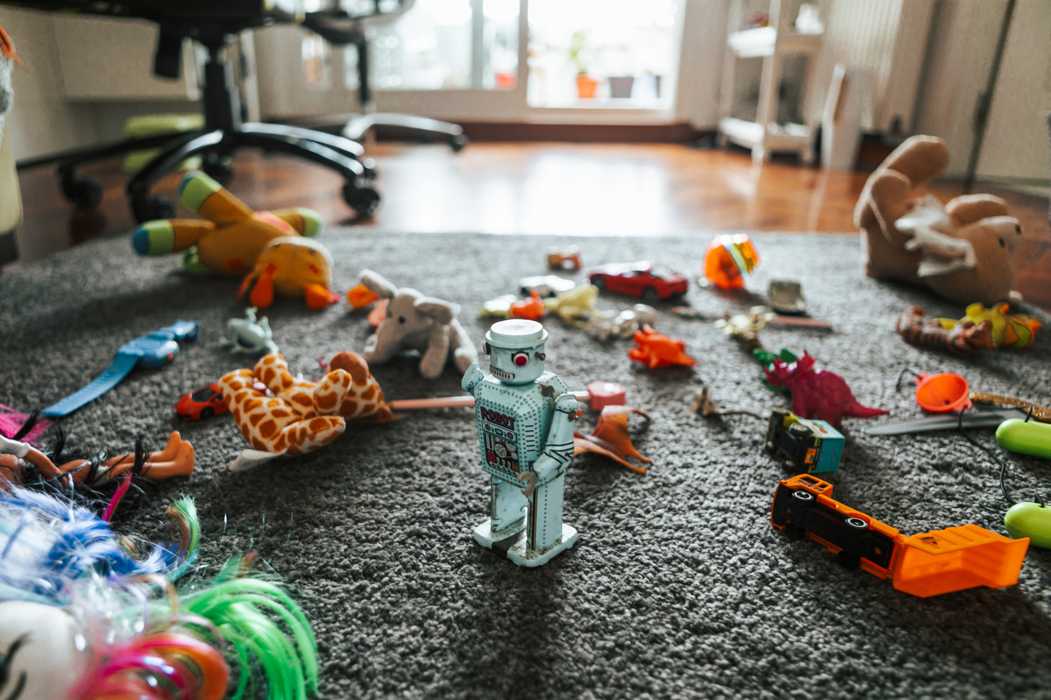 A cluttered playroom floor is strewn with various toys, including a toy robot, a giraffe, and multiple stuffed animals and vehicles