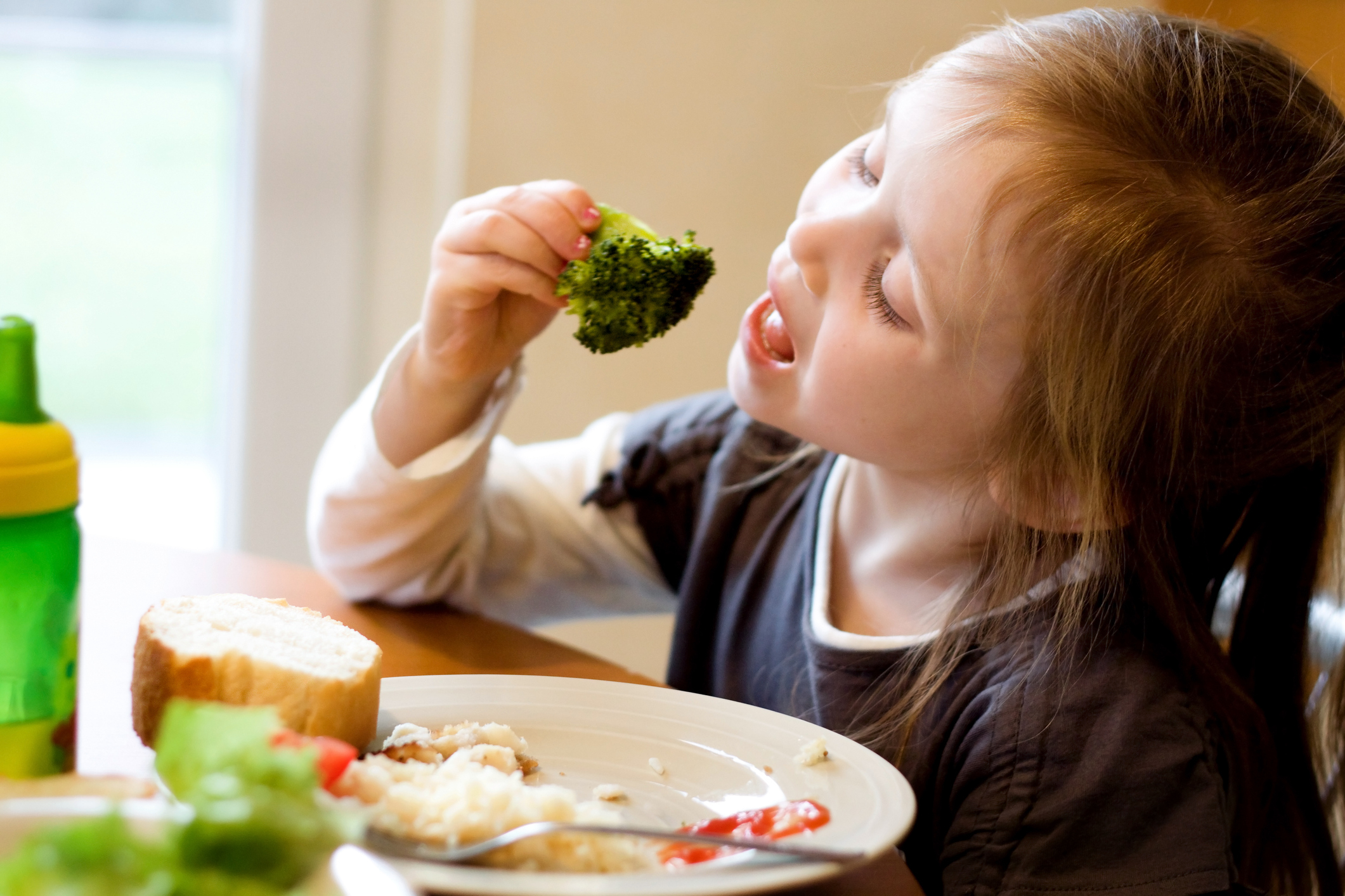 A young girl eats broccoli at a dining table with a plate of food, including bread and rice