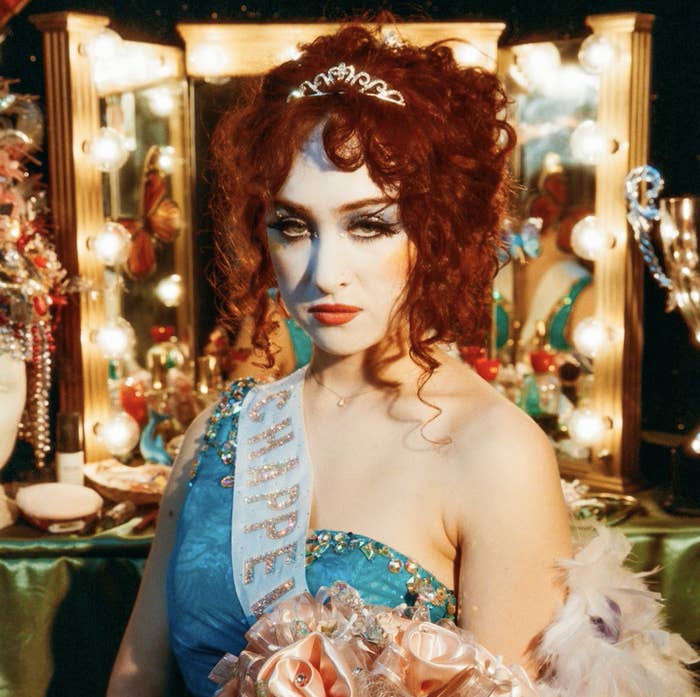 Chappell Roan with curly hair, wearing a tiara and a decorated sash, stands in front of a vanity filled with lights and makeup items, holding a bouquet of flowers