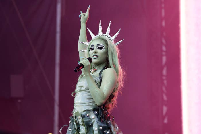 Chappell Roan performing on stage wearing a metallic outfit with spikes and a crown resembling the Statue of Liberty, holding a microphone with one hand raised