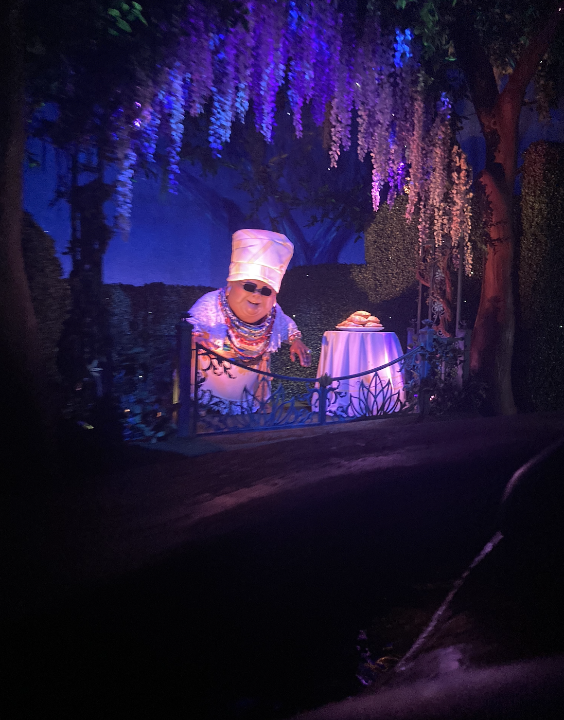 Chef Gusteau from Ratatouille is joyfully leaning over a table in a scenic, whimsical outdoor setting with hanging flowers