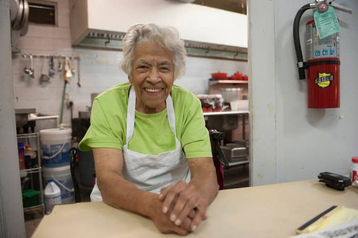 An elderly woman with white hair smiles warmly while leaning on a kitchen counter. She is wearing a green shirt and a white apron. Kitchen equipment is visible in the background