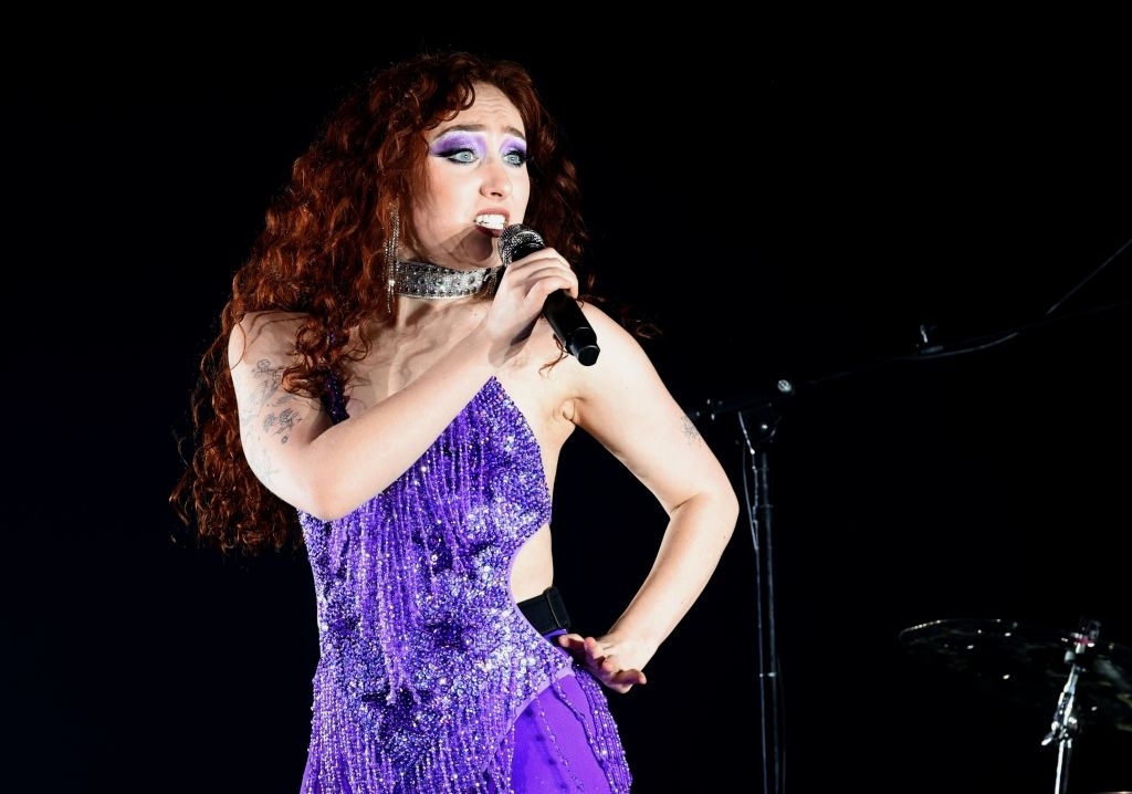 Chappell Roan with long, curly red hair performing on stage in a sparkling, sleeveless, purple dress with a large silver choker necklace