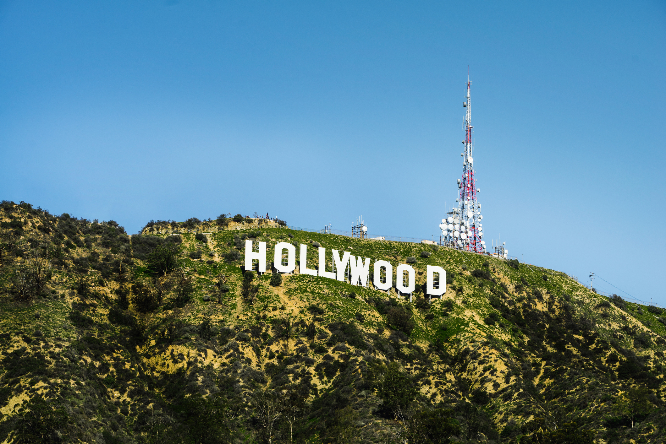 The image shows the iconic Hollywood sign on a hill, with a communication tower in the background, under a clear sky