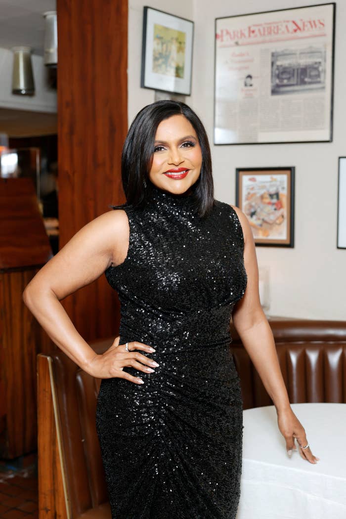 Mindy Kaling, wearing a sleeveless, sequined dress, poses with a hand on her hip in a restaurant setting with framed articles and pictures on the wall behind her