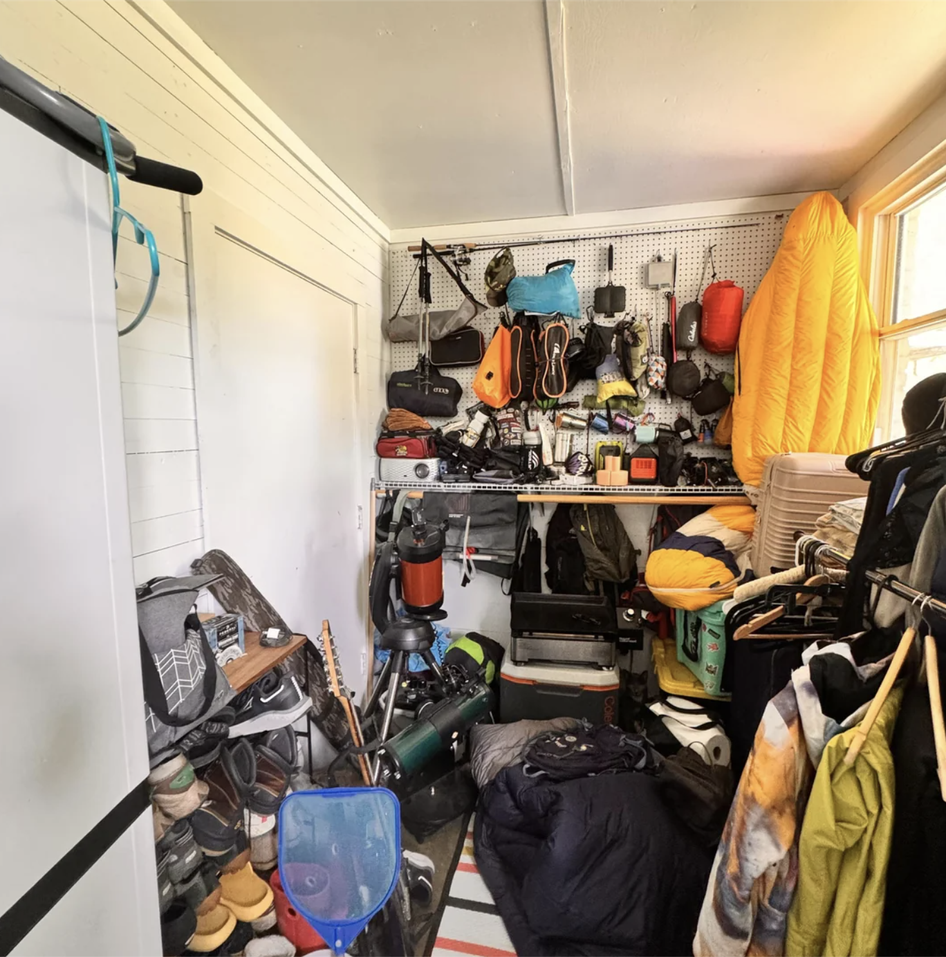 A cluttered storage room filled with outdoor gear, tools, and miscellaneous items. A bright jacket is hanging on the wall. Shelves are stacked with various equipment