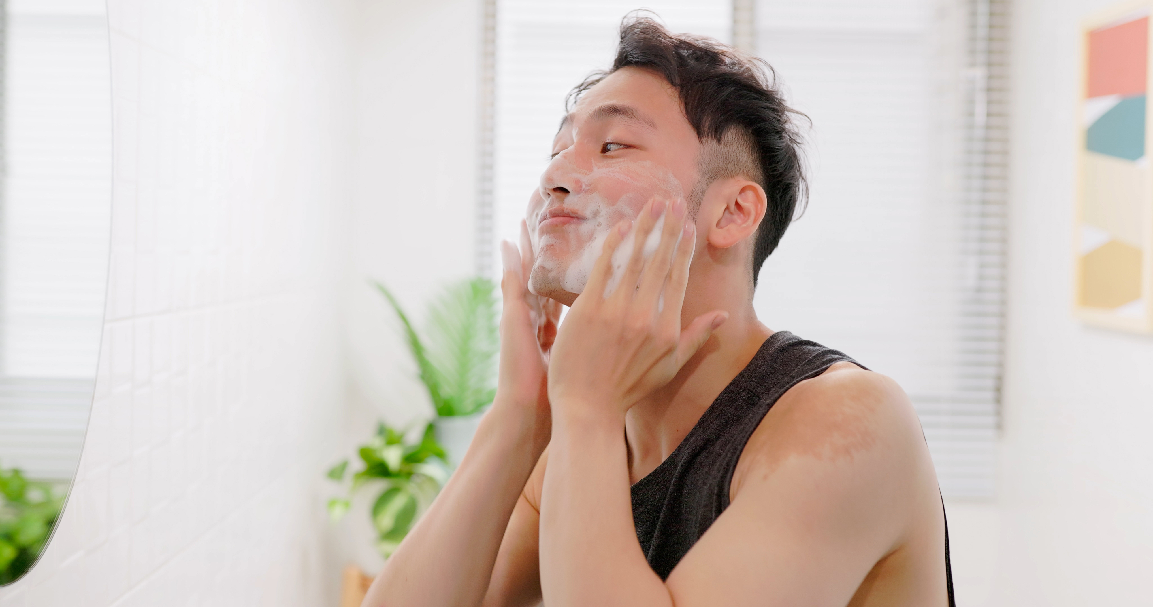 A person with short hair is cleansing their face with a foamy cleanser in a bright bathroom, standing in front of a mirror