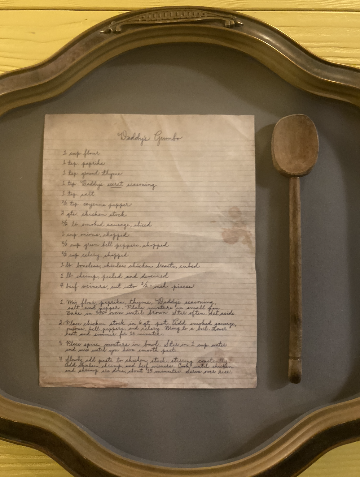 A framed handwritten recipe for &quot;Daddy&#x27;s Gumbo&quot; is displayed next to a wooden spoon. The recipe includes ingredients and cooking instructions