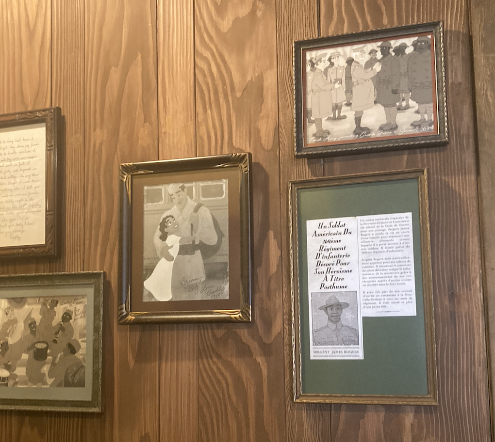 Four framed items on a wooden wall, including a portrait of a woman in military uniform, a newspaper clipping about T/Sgt. John Robert Slater, and illustrations