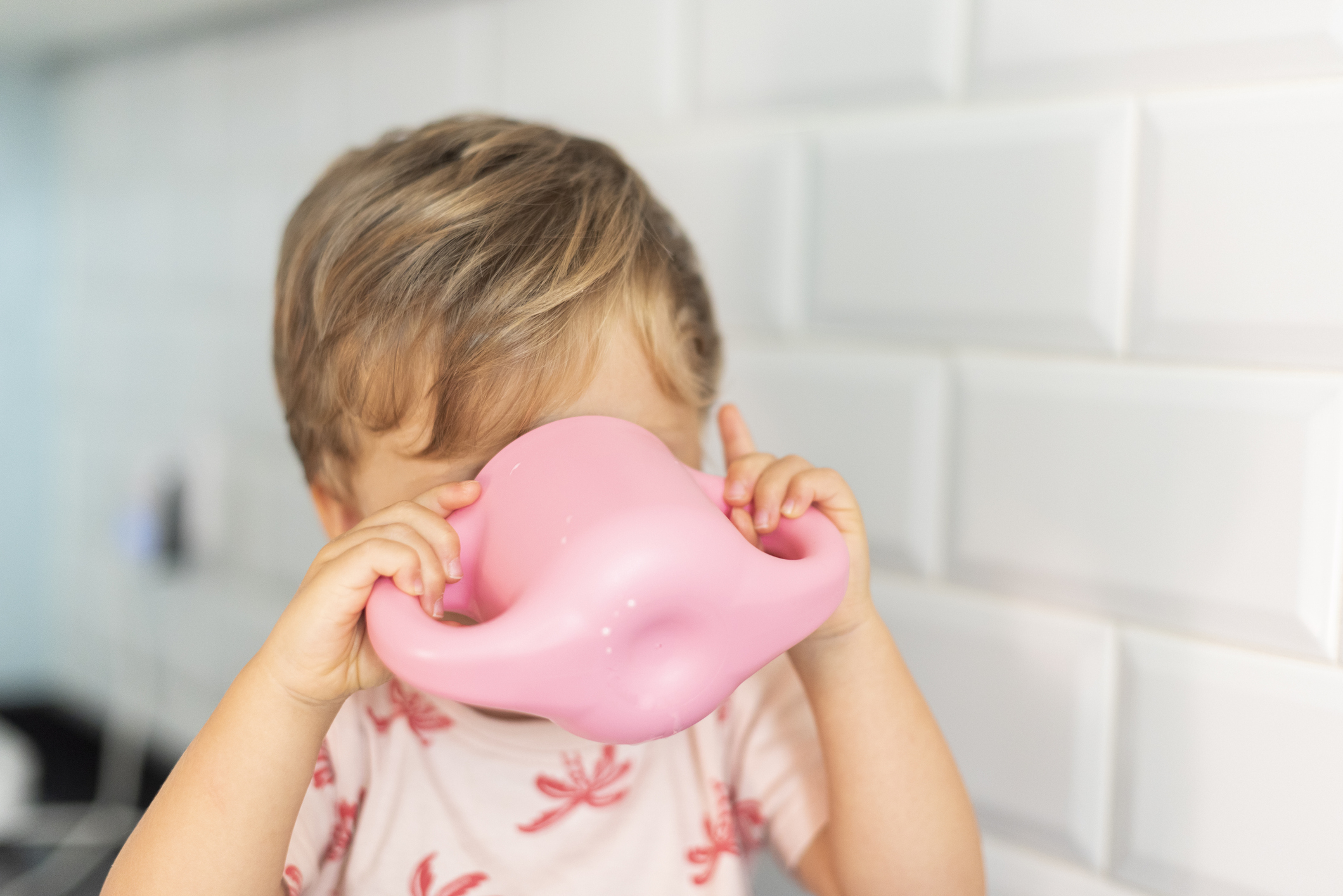 Toddler drinking from a pink mustache-shaped cup with a tiled wall in the background