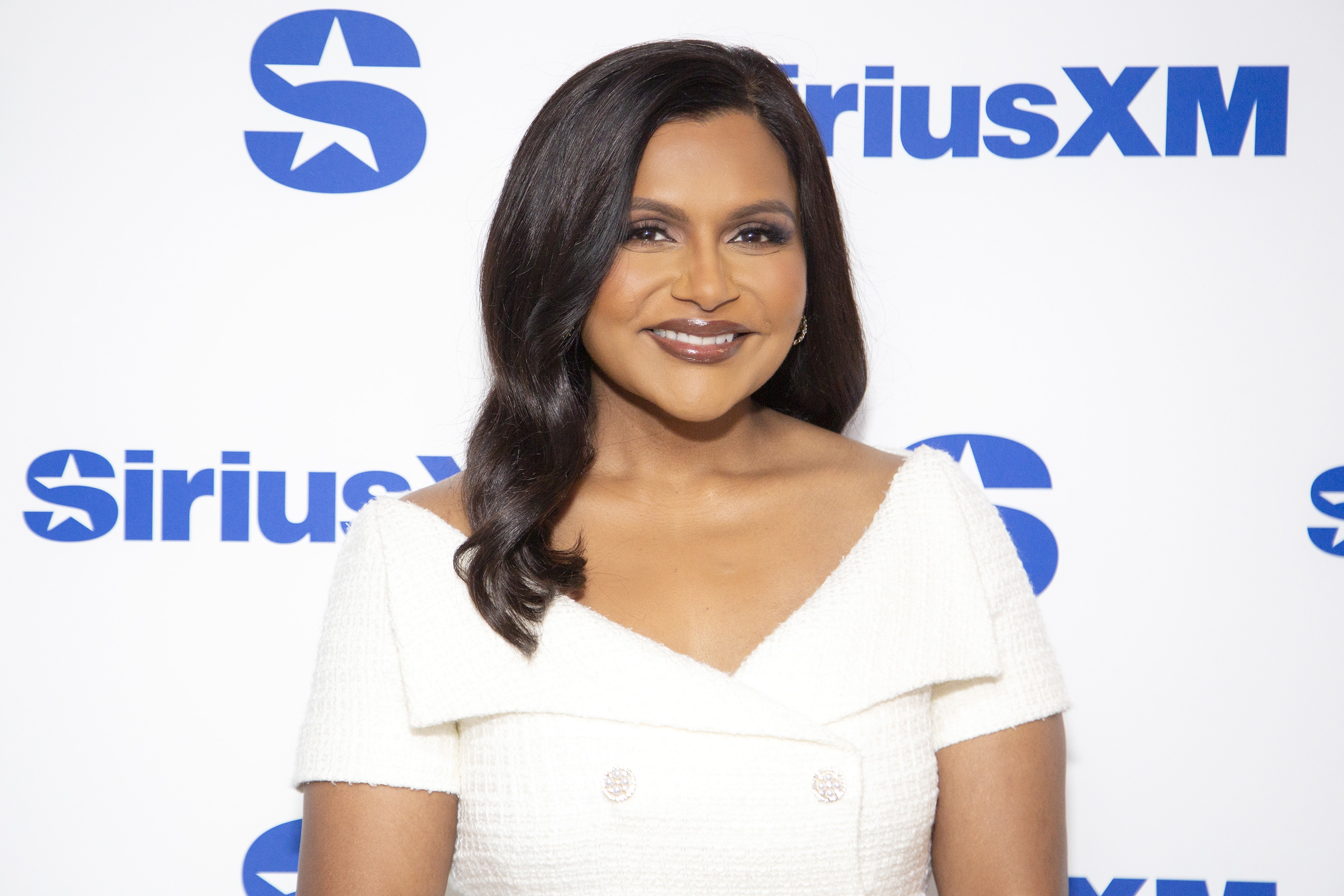 Mindy Kaling poses in front of a SiriusXM backdrop, wearing an elegant off-the-shoulder dress with a confident smile