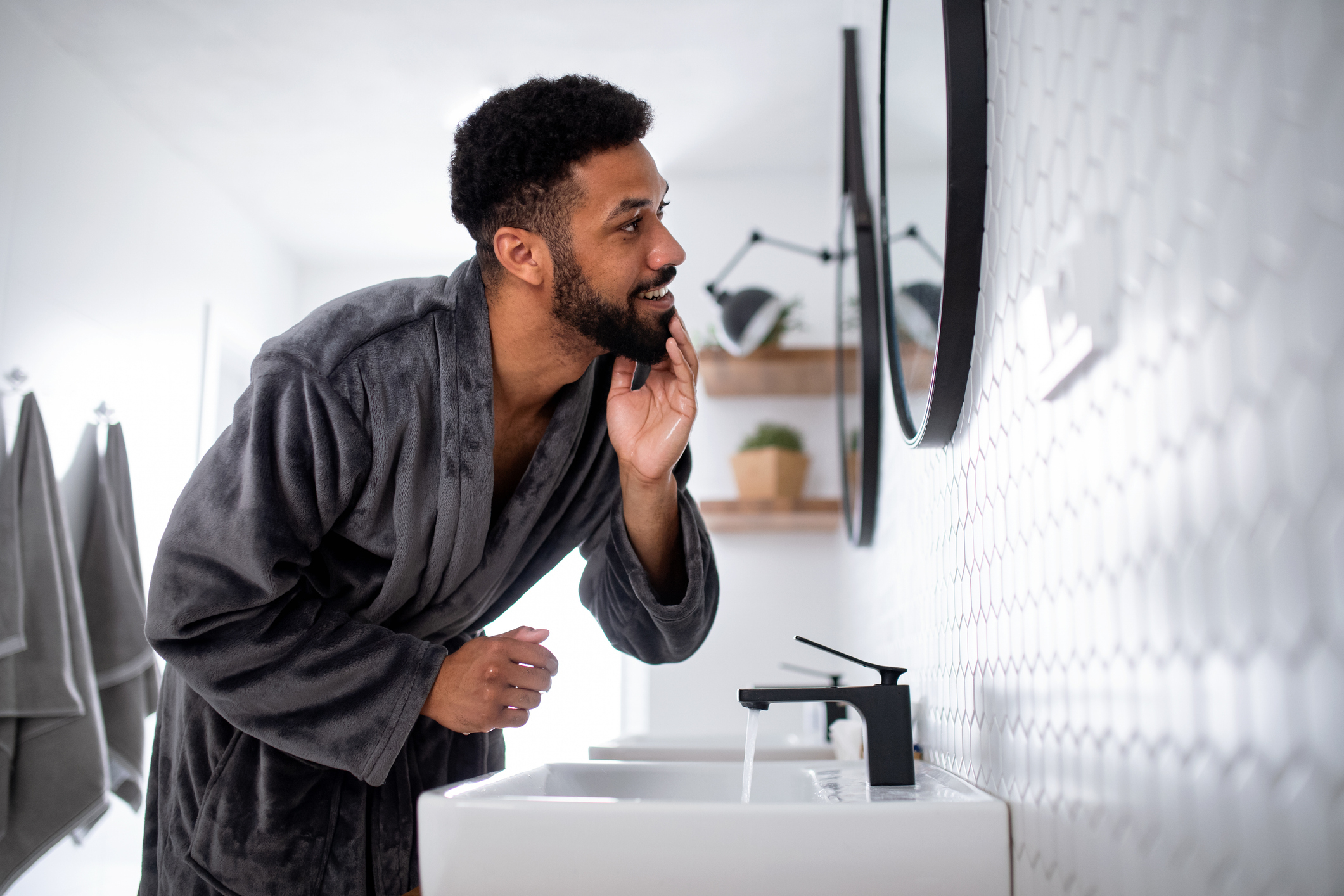 A man in a robe examines his beard in a bathroom mirror