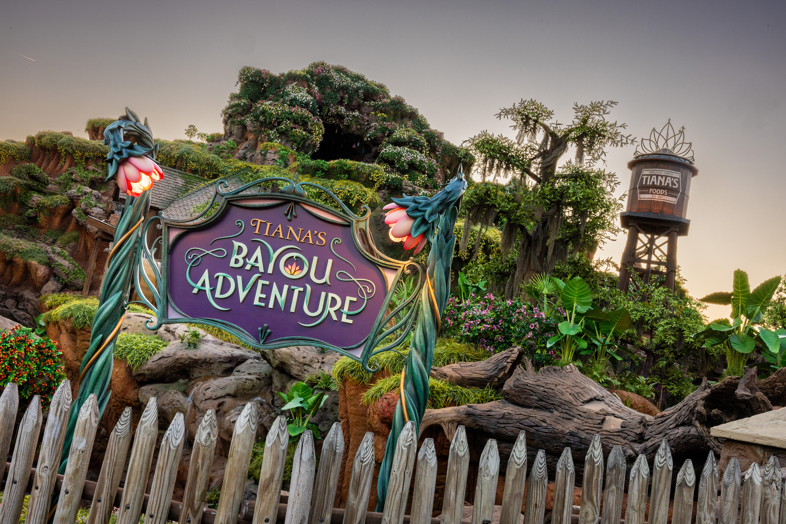 Tiana&#x27;s Bayou Adventure sign in front of a lush, green landscape with trees, flowers, and a water tower in the background