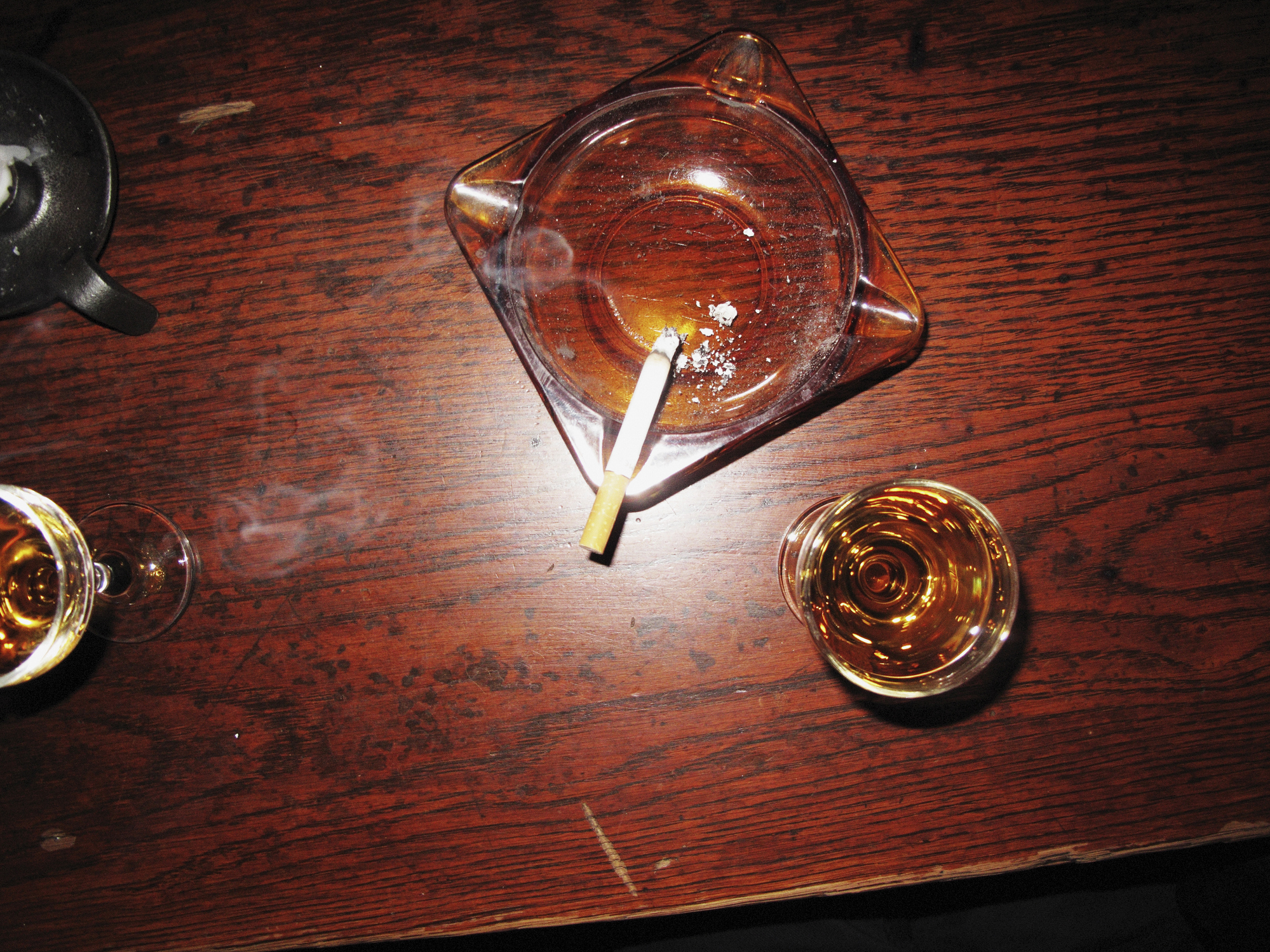 A cigarette burns in an ashtray on a wooden table with two glasses containing amber liquid