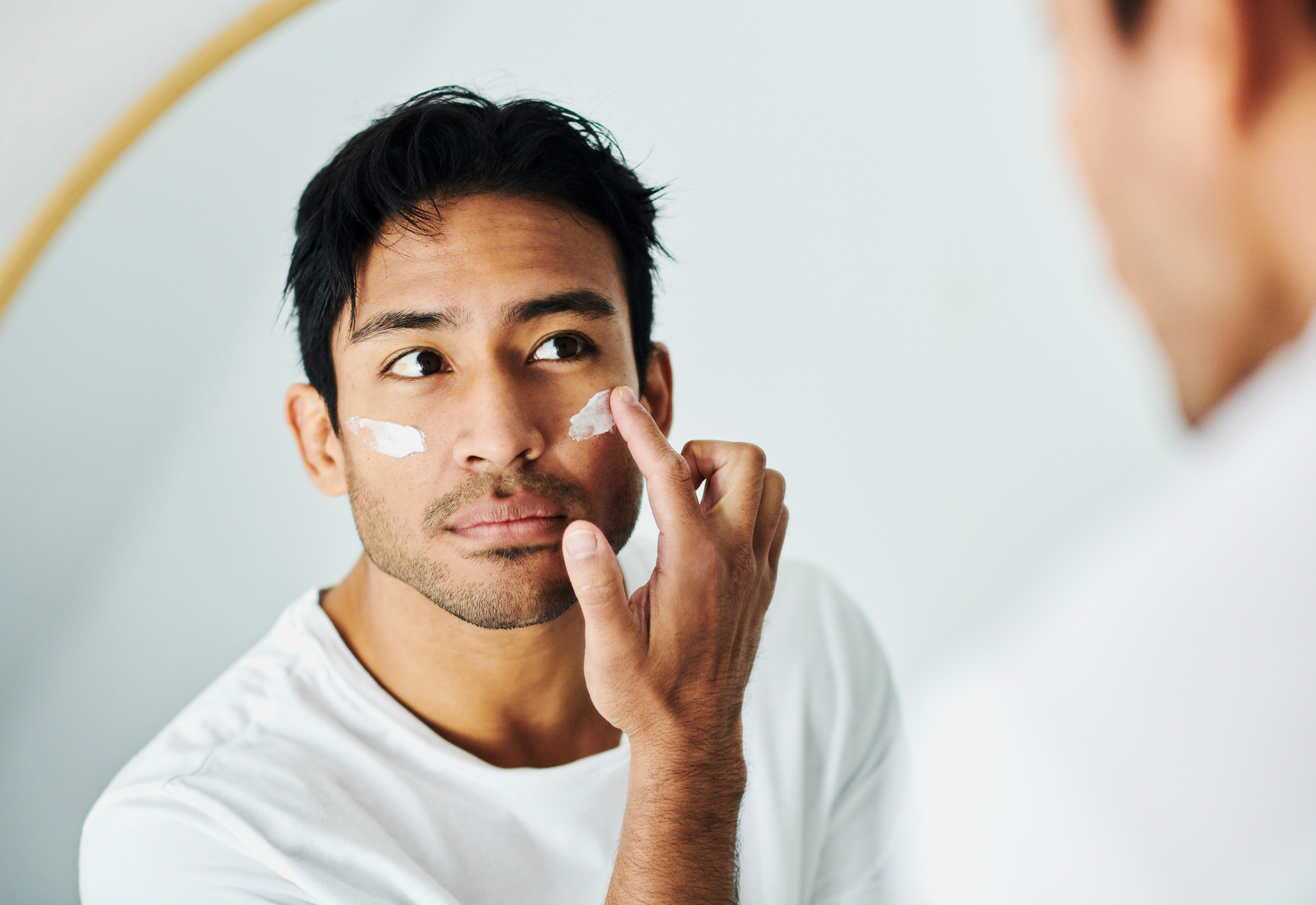 A man in a white shirt applies skincare cream to his face while looking at himself in a bathroom mirror