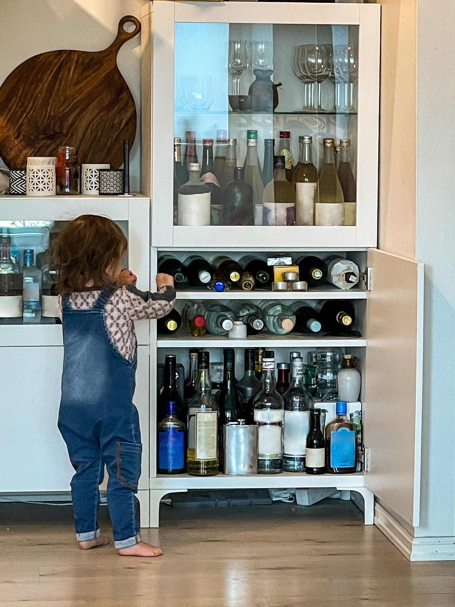 A toddler in overalls is reaching into a cabinet filled with various bottles and glasses