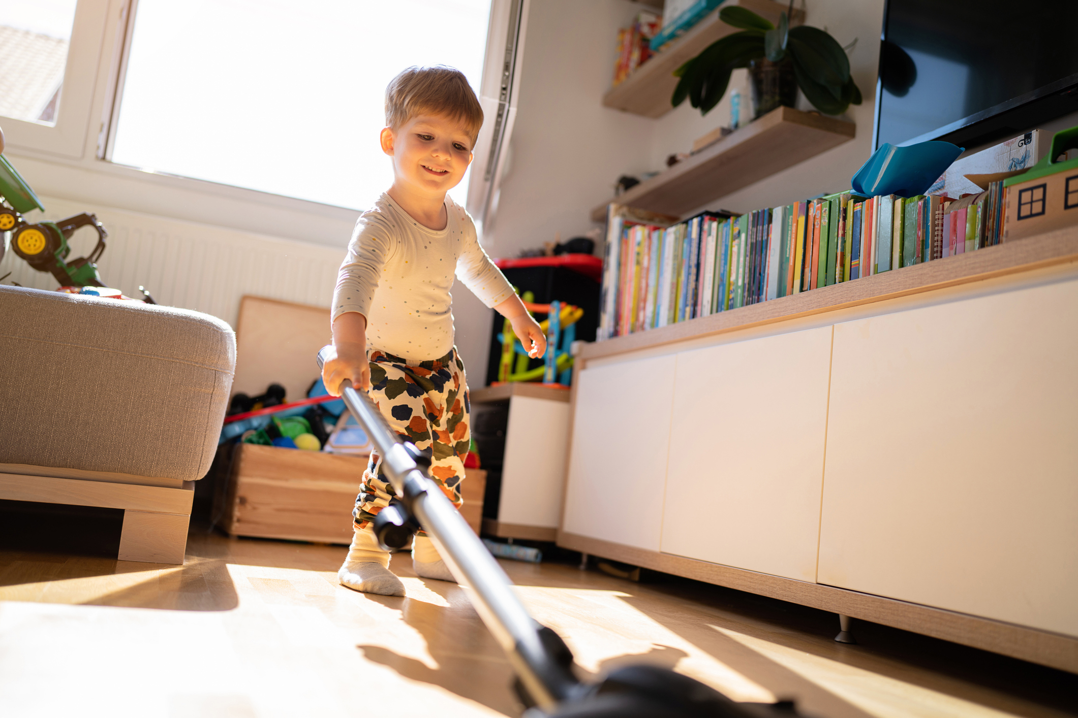 Young child smiling while vacuuming a living room filled with toys and books