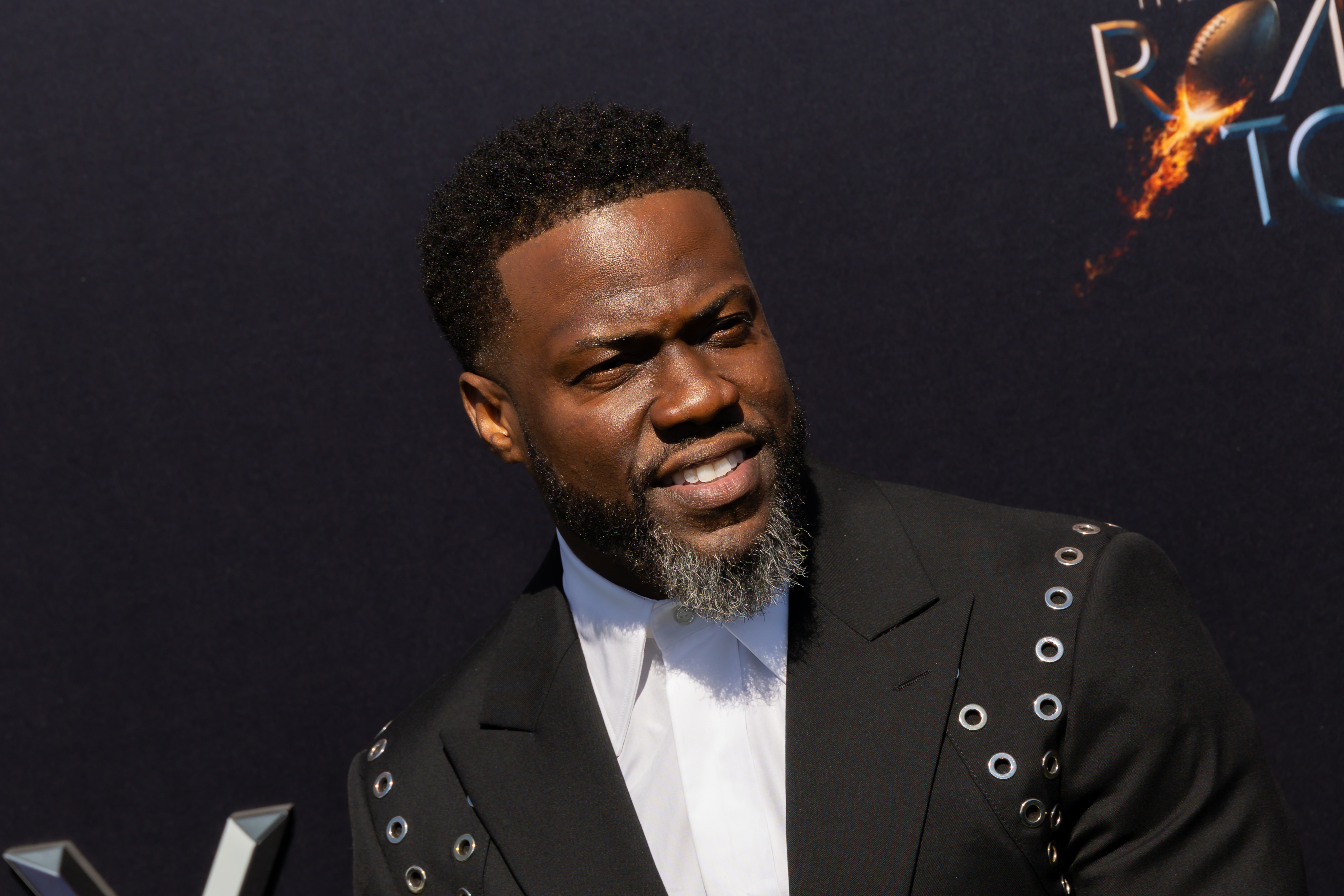 Kevin Hart wearing a stylish black suit with grommet detailing at the premiere event of &quot;The Road to&quot; against a dark backdrop