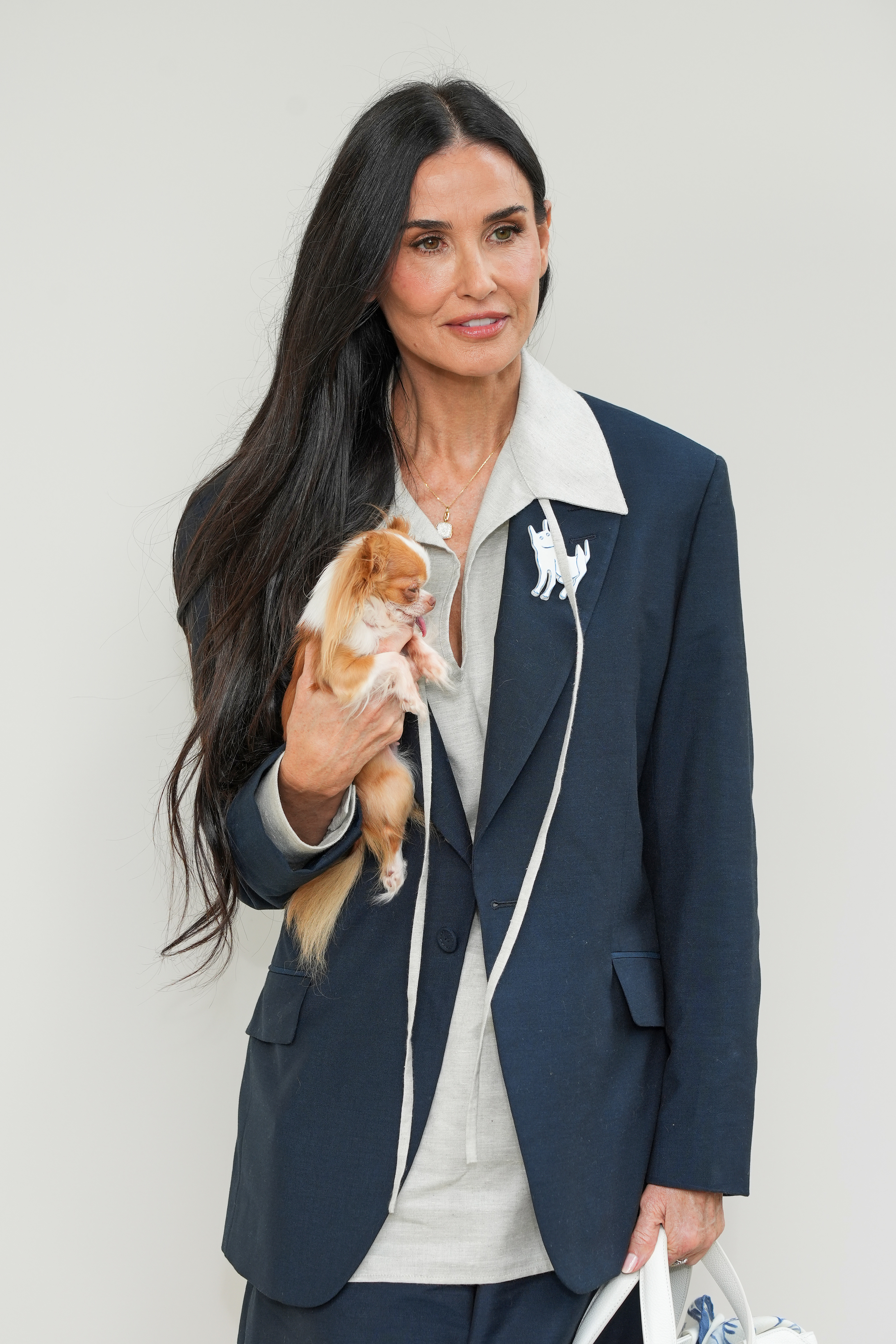 Demi Moore holding a small dog, wearing a long dark blazer over a light outfit, standing against a plain background