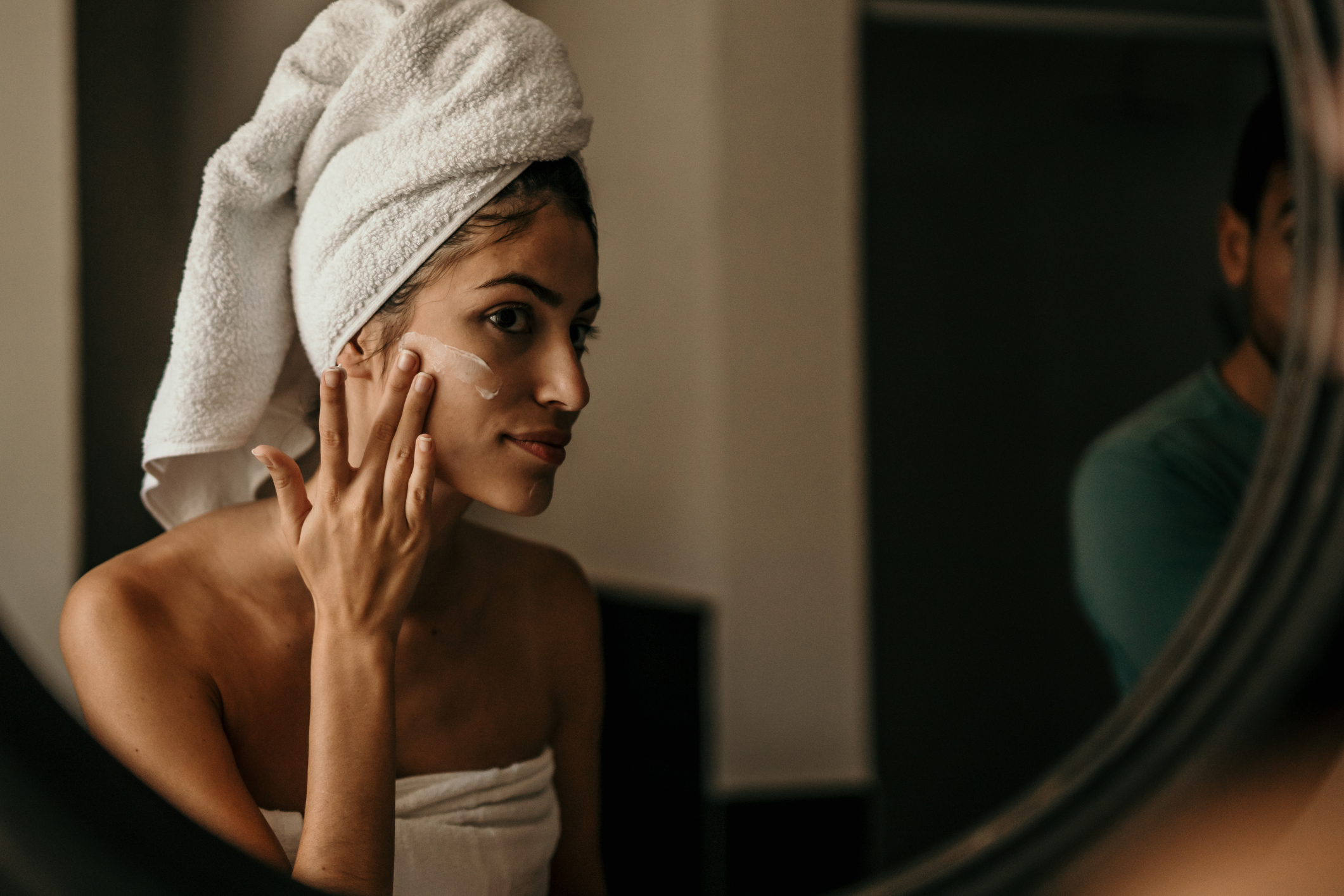 A woman wrapped in a towel and with a towel on her head, applies cream to her face, reflecting a skincare routine. Another person is partly visible in the background