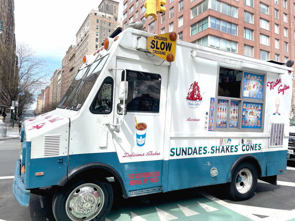 An ice cream truck parked on a city street with a sign reading &quot;Children Slow Crossing&quot; on top. The truck offers sundaes, shakes, and cones