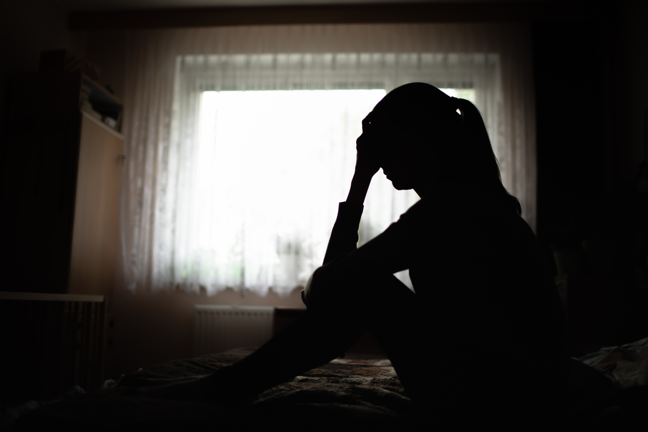 Silhouette of a person sitting with their hand on their forehead in front of a window with sheer curtains