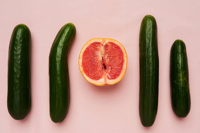 Four cucumbers surround half of a grapefruit placed on a light pink surface in a vertical alignment