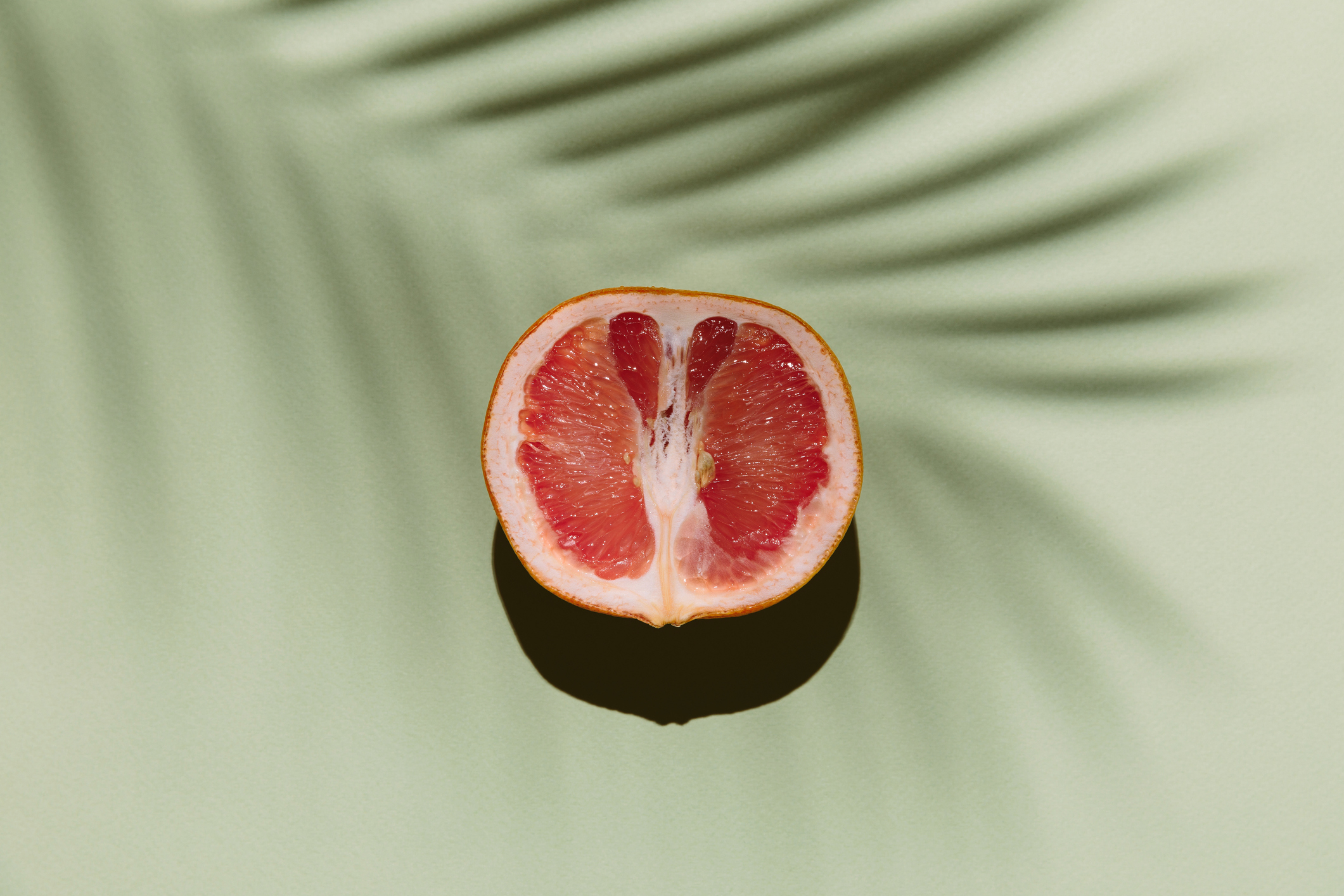A halved grapefruit casting a shadow on a smooth surface with a palm leaf shadow in the background