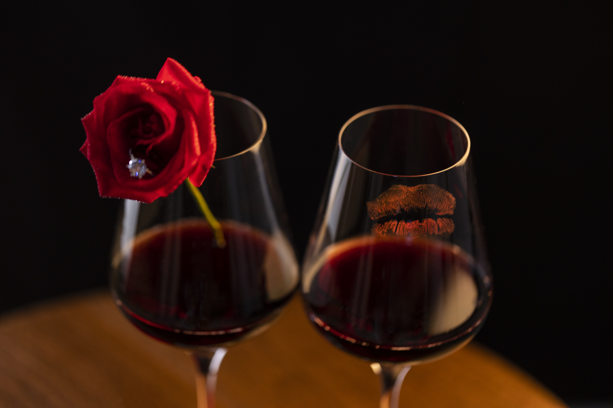 Two wine glasses filled with red wine; one has a red rose resting on it, the other has lipstick marks on its rim