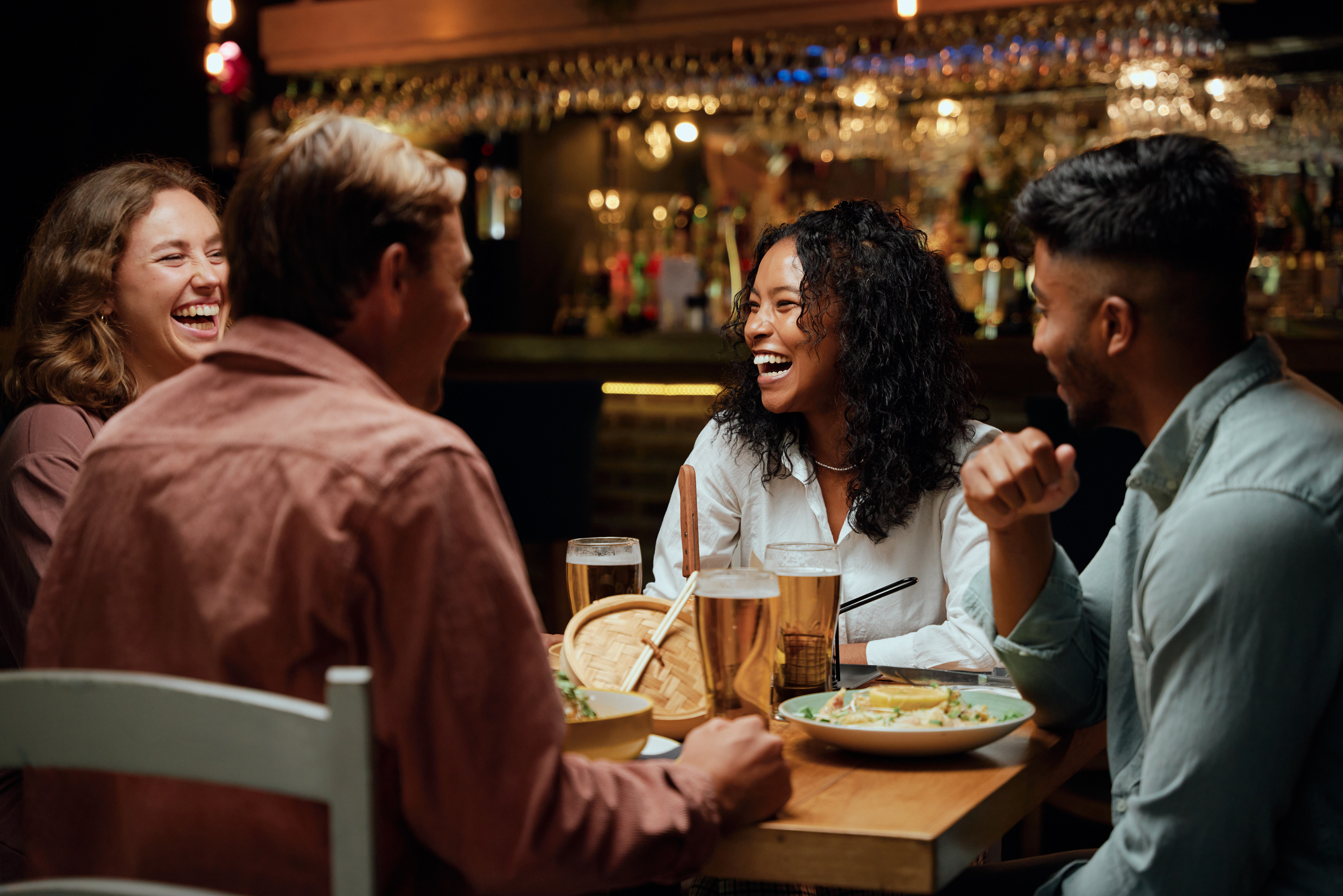 Four people share a meal at a restaurant, smiling and engaged in conversation, with plates of food and glasses of beer on the table