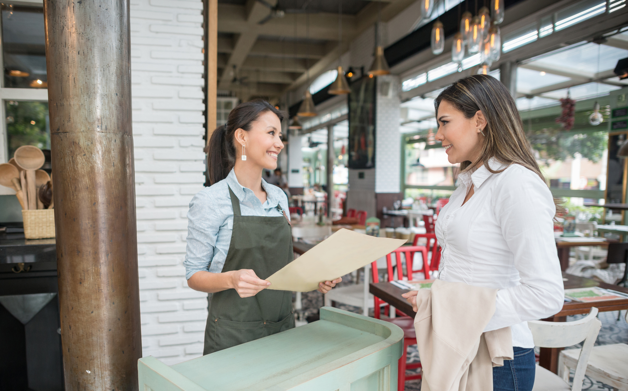 A hostess holding a menu stands behind a counter in a restaurant, conversing and smiling with a female customer dressed in a white blouse