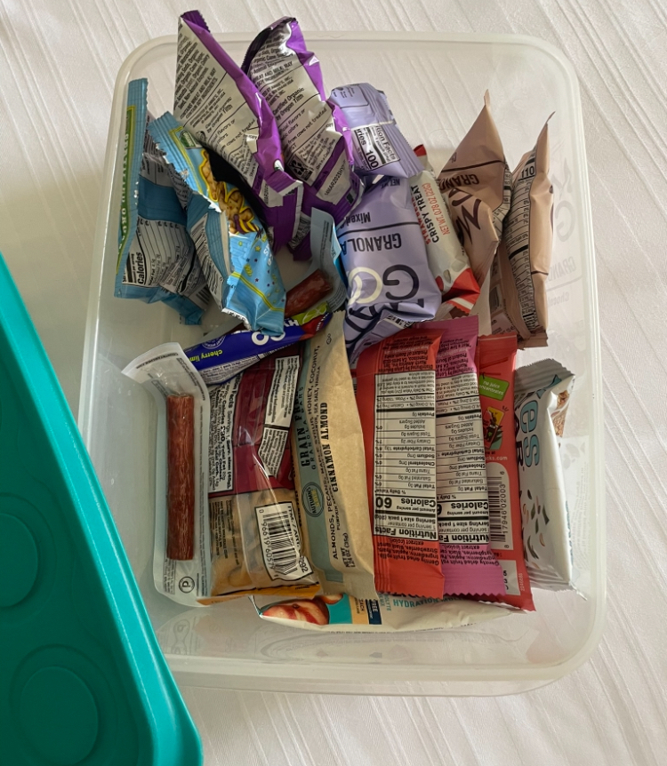 An open plastic container filled with various individually wrapped snacks and bars, some nutritional information visible on the packaging