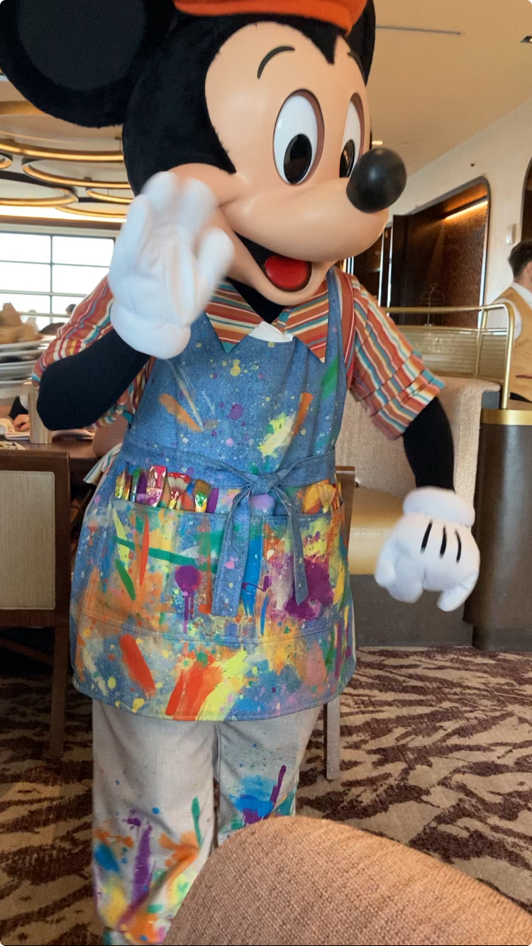 Mickey Mouse wears a colorful apron splattered with paint and waves indoors