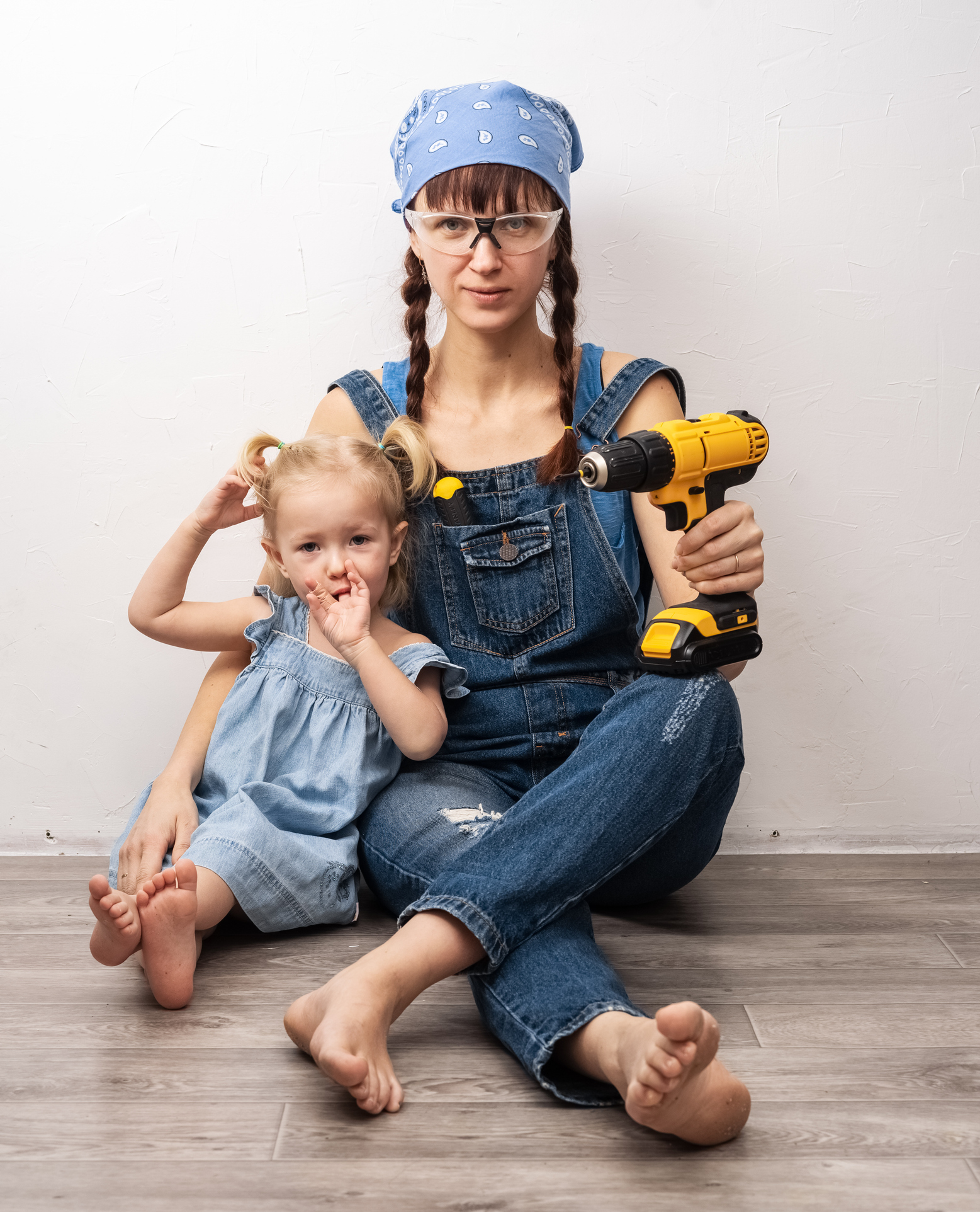 A woman wearing overalls and a bandana, seated on the floor holding a power drill, sits next to a young girl in a blue dress with pigtails