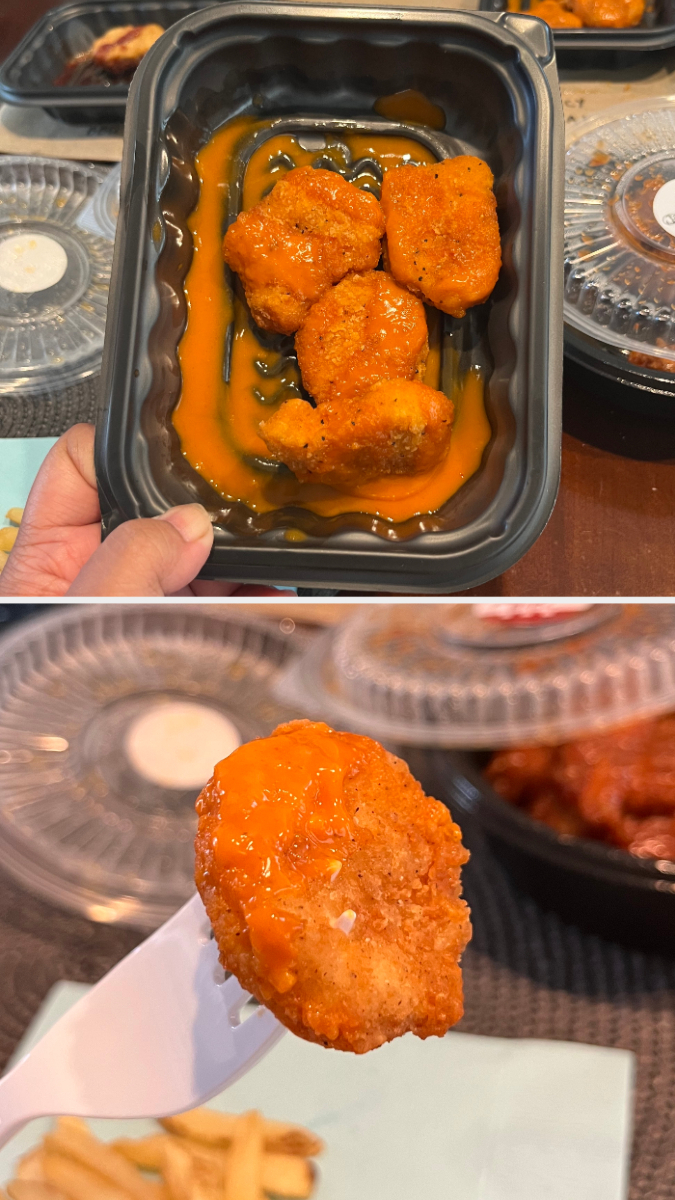 Close-up of boneless buffalo chicken wings in a black plastic container, with one being held up on a fork, fries in the background