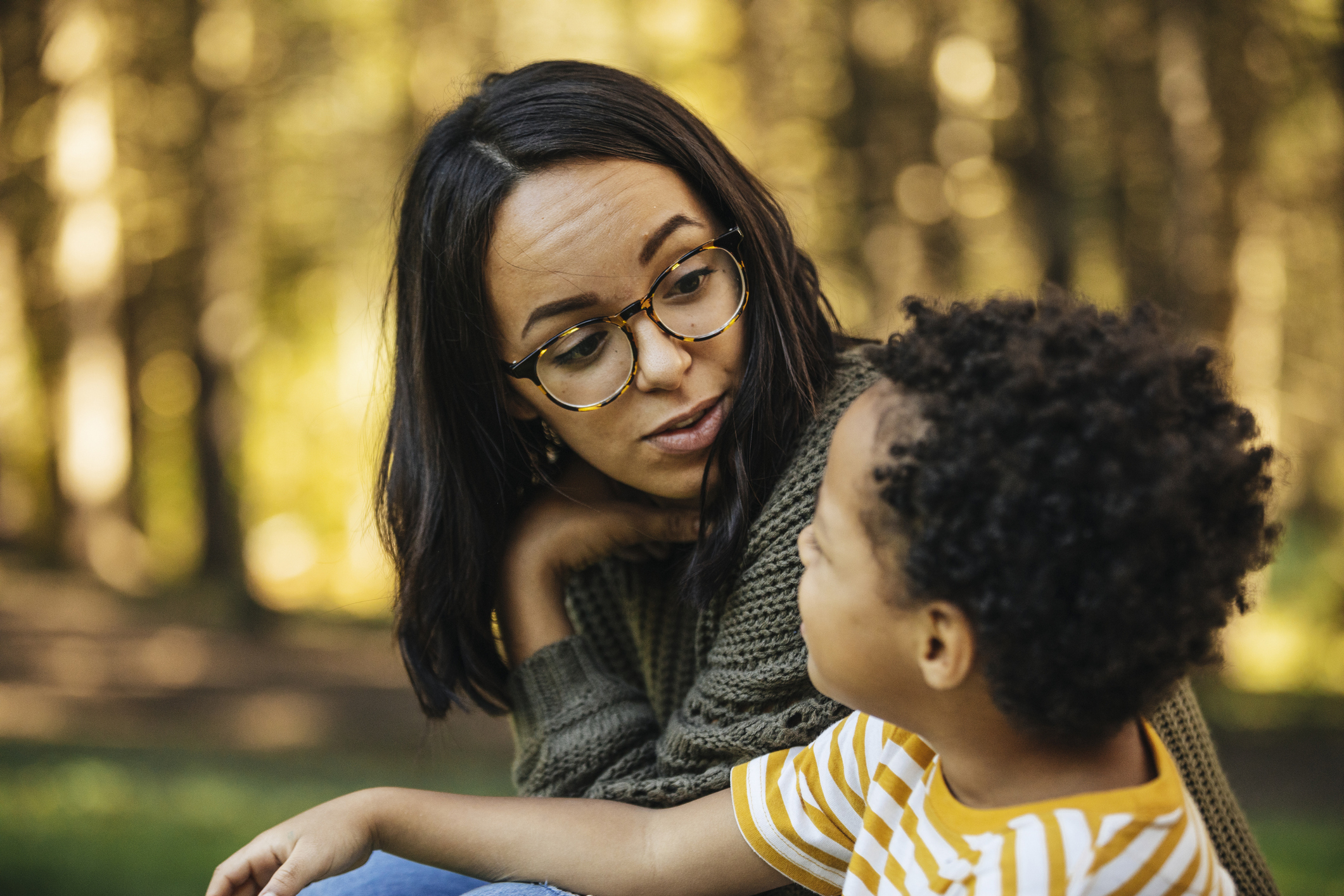 A woman wearing glasses and a child in a striped shirt are sitting outdoors, engaged in conversation