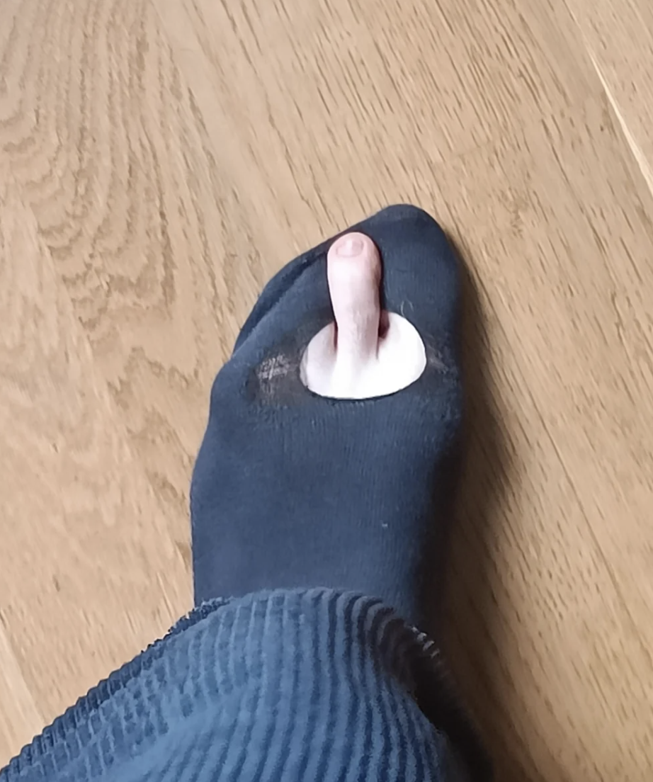 A person&#x27;s foot in a black sock with a hole, revealing their big toe, standing on a wooden floor