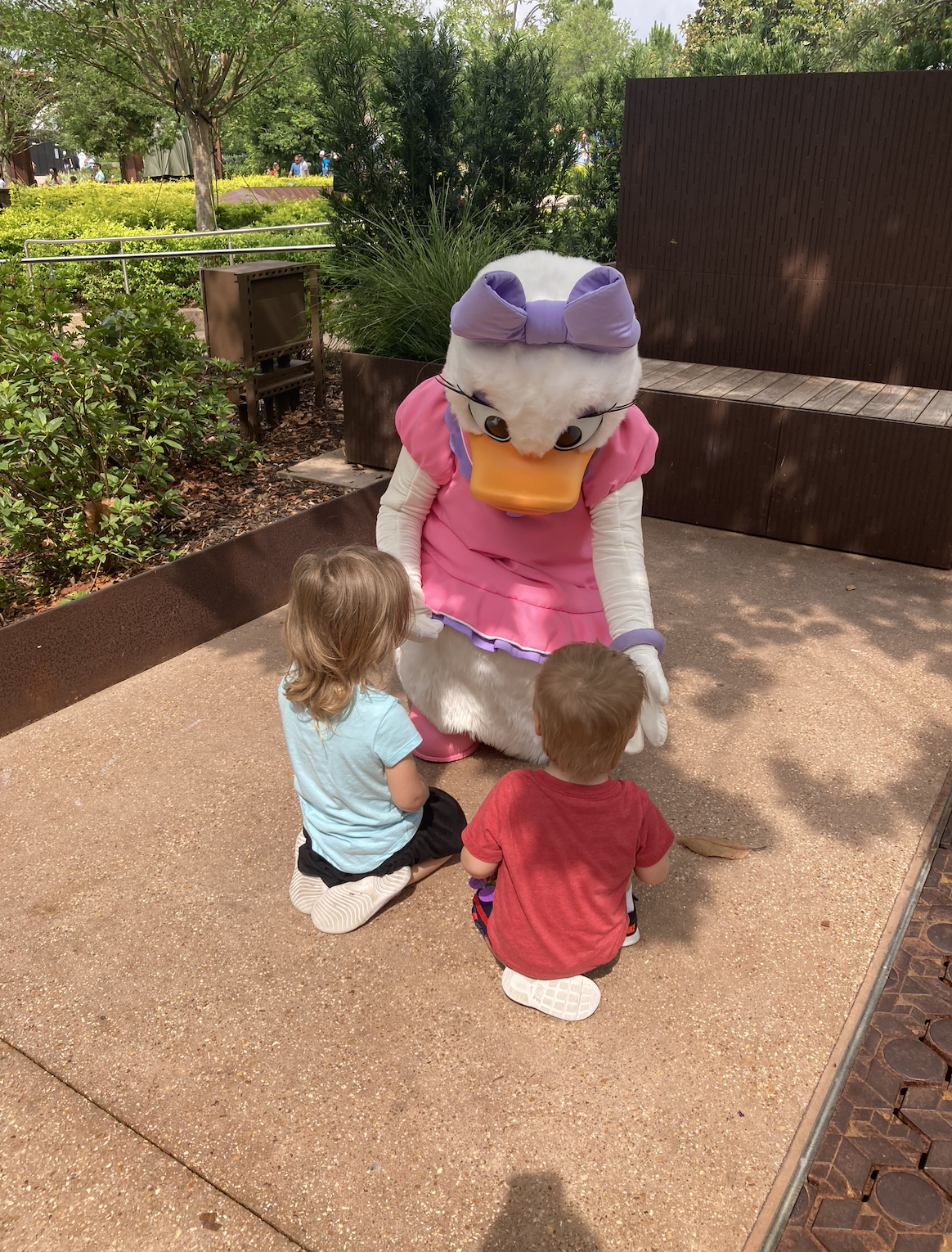 Two children are kneeling on the ground, facing Daisy Duck, who is dressed in a pink dress and purple bow, in an outdoor setting