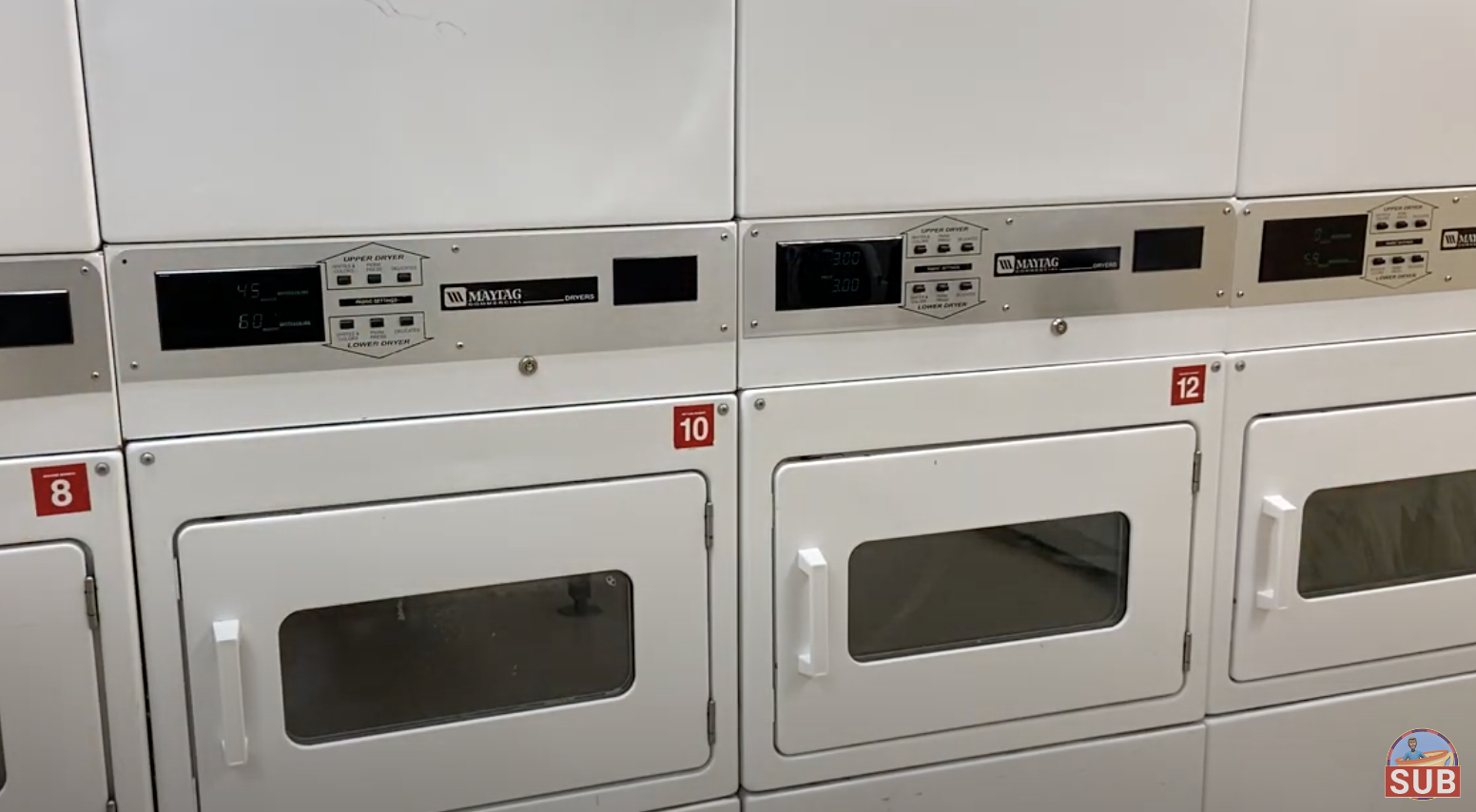 Four white commercial dryers in a laundromat are stacked two-high. Doors are closed. A small logo &quot;SUB&quot; is seen in the bottom right corner