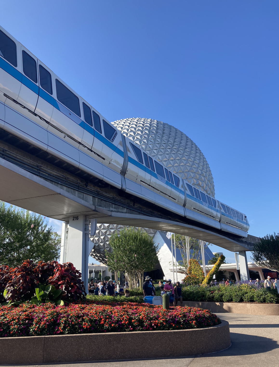 Monorail passes by Epcot&#x27;s Spaceship Earth with vibrant flowers and visitors in the foreground