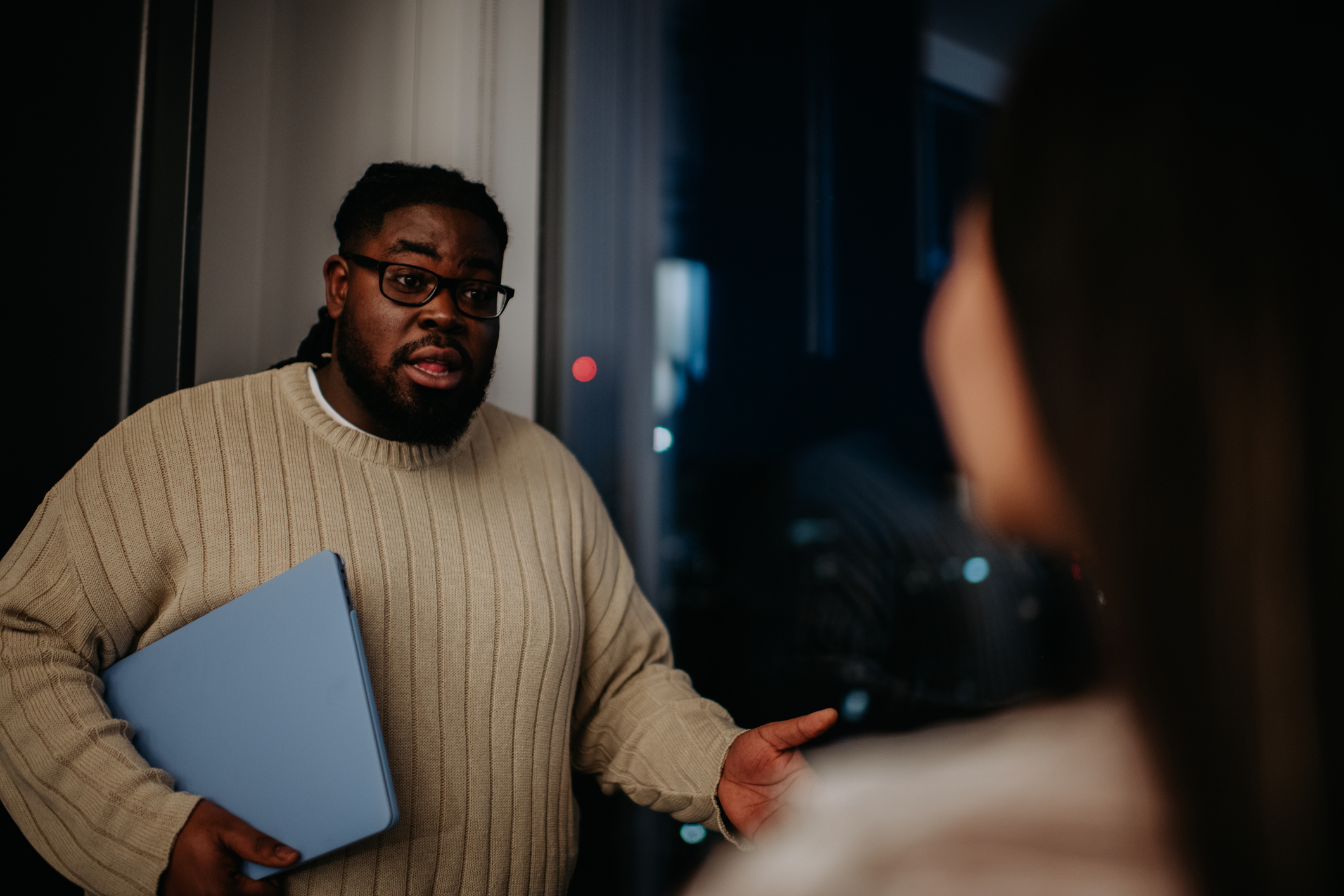 A man wearing a sweater and carrying a laptop is engaged in a conversation with a woman, seen from behind. The setting appears to be a professional environment